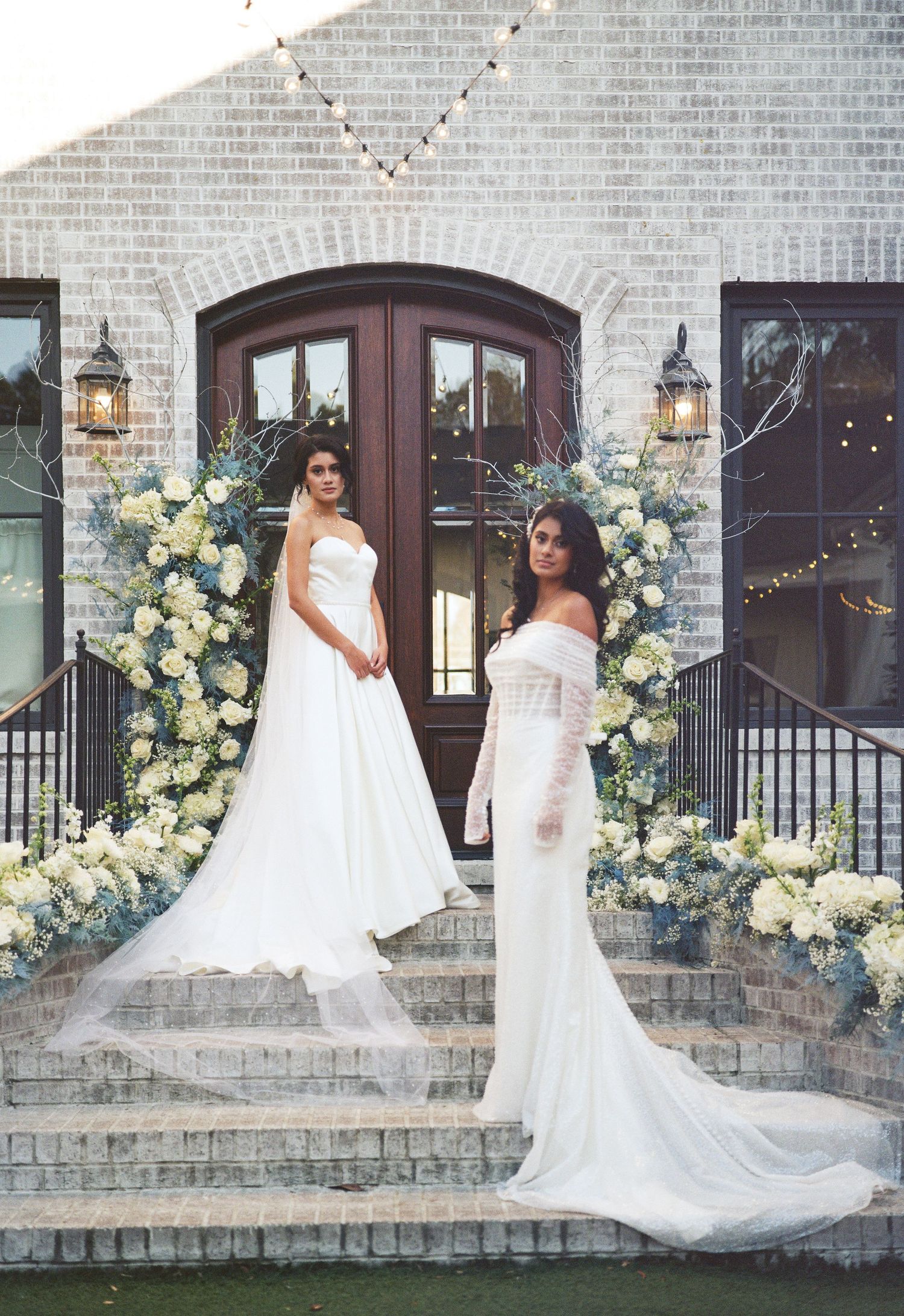 Two brides in white wedding gowns pose on stone steps decorated with yellow floral arrangements.