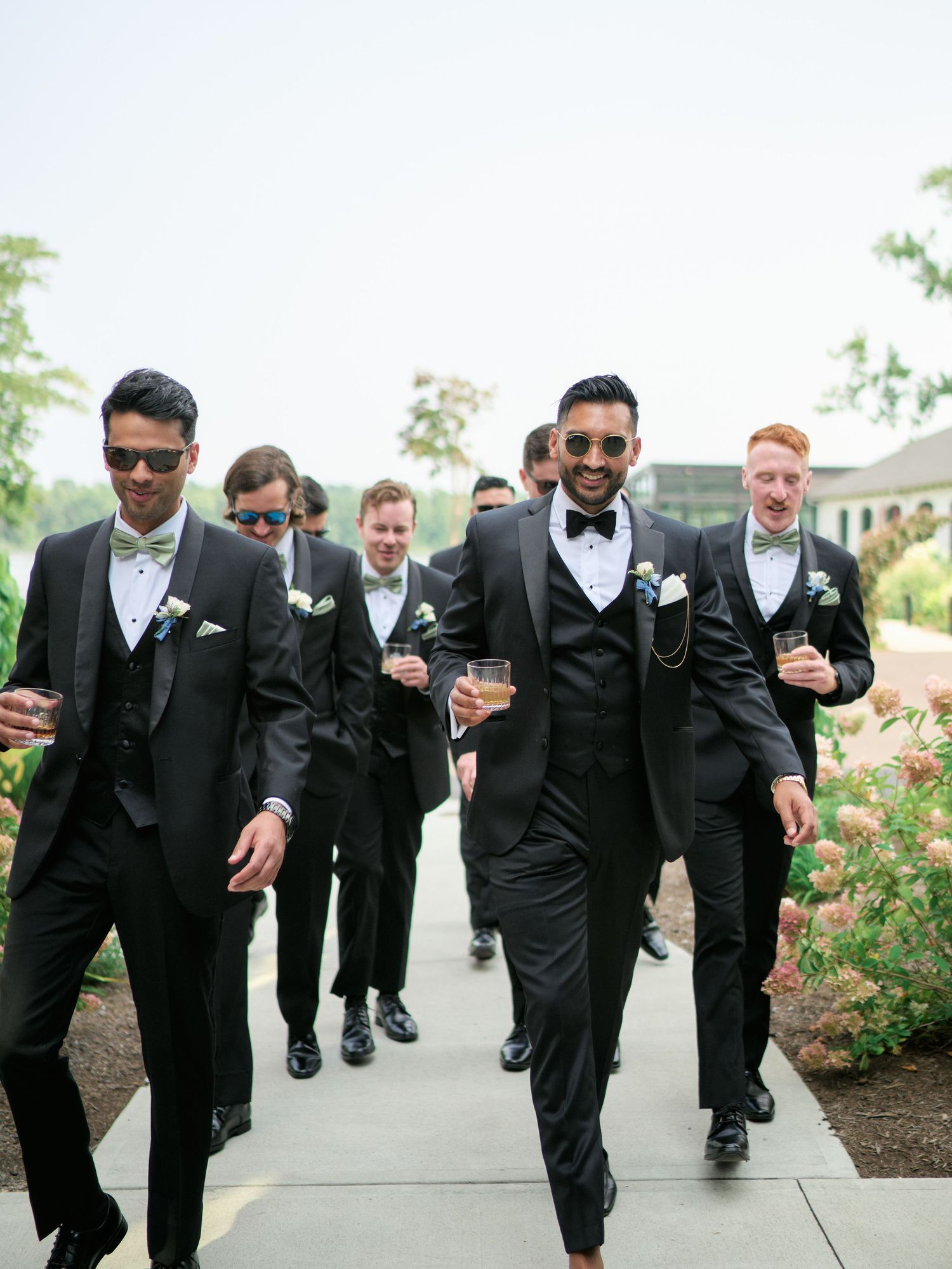 Groomsmen walking in a line outdoors wearing matching black tuxedos with sunglasses and bow ties on a wedding day.