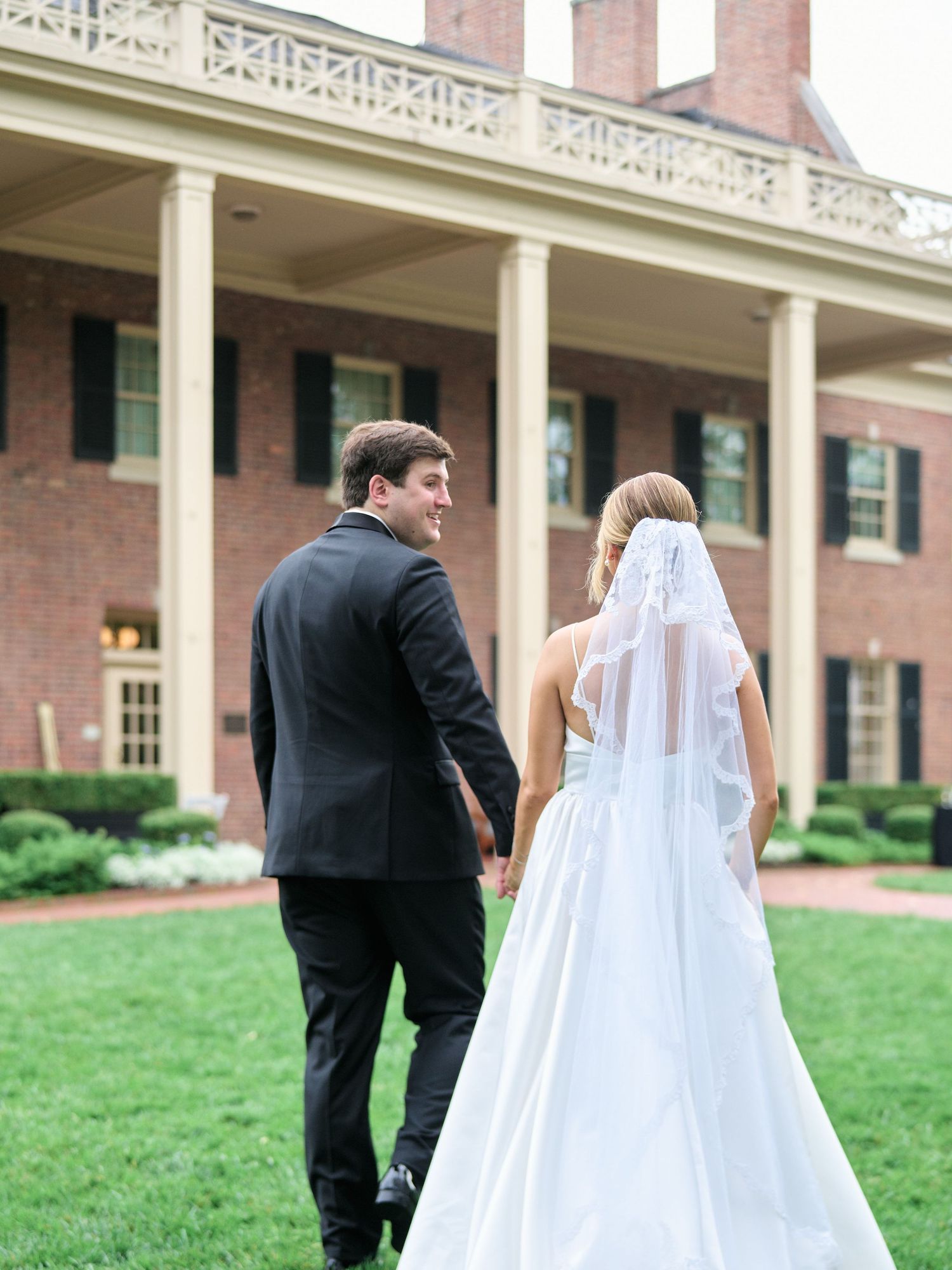 Wedding couple walking away from camera toward a grand colonial style mansion.
