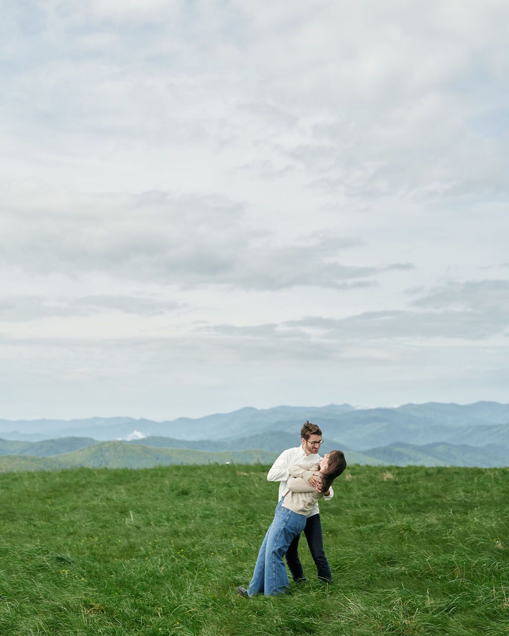A couple embraces in a romantic moment on a grassy hilltop overlooking distant blue mountain ranges.