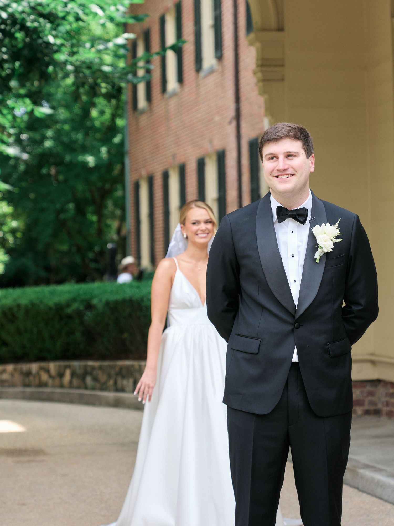 A couple in formal wedding attire poses together under a columned archway near a brick building.