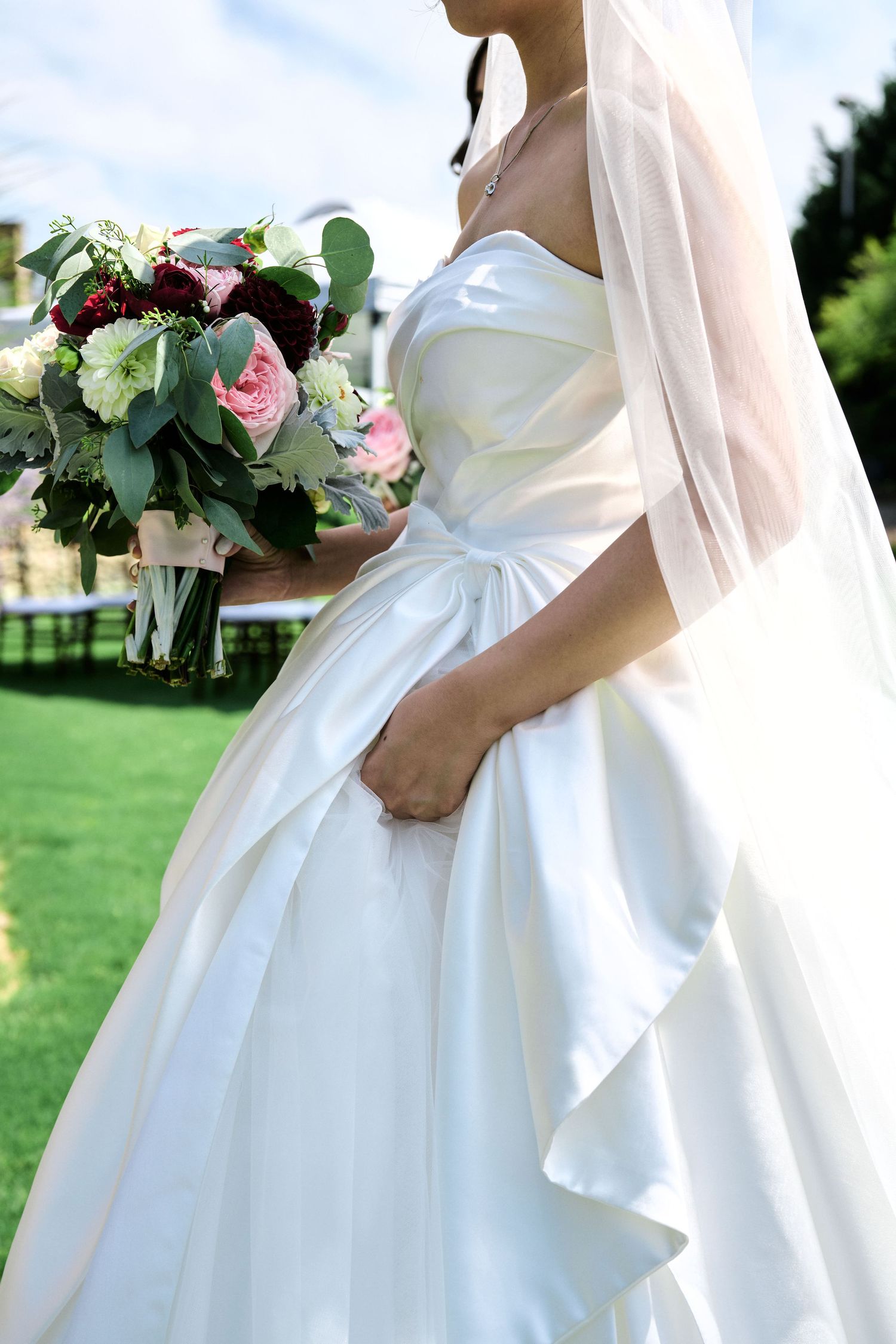 Close-up detail of a white wedding dress and bouquet featuring pink roses and greenery.