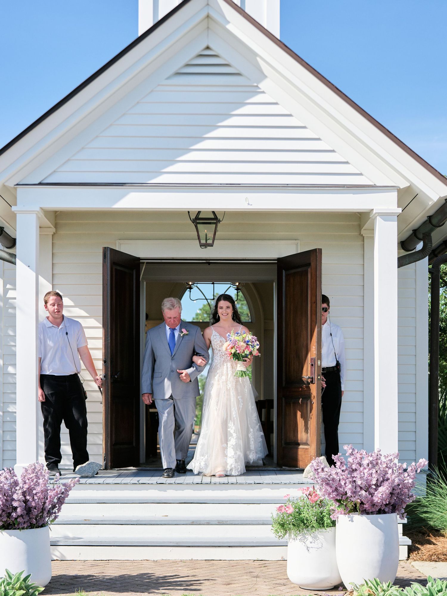 A small white chapel with potted purple flowers flanking the entrance steps on a sunny day.