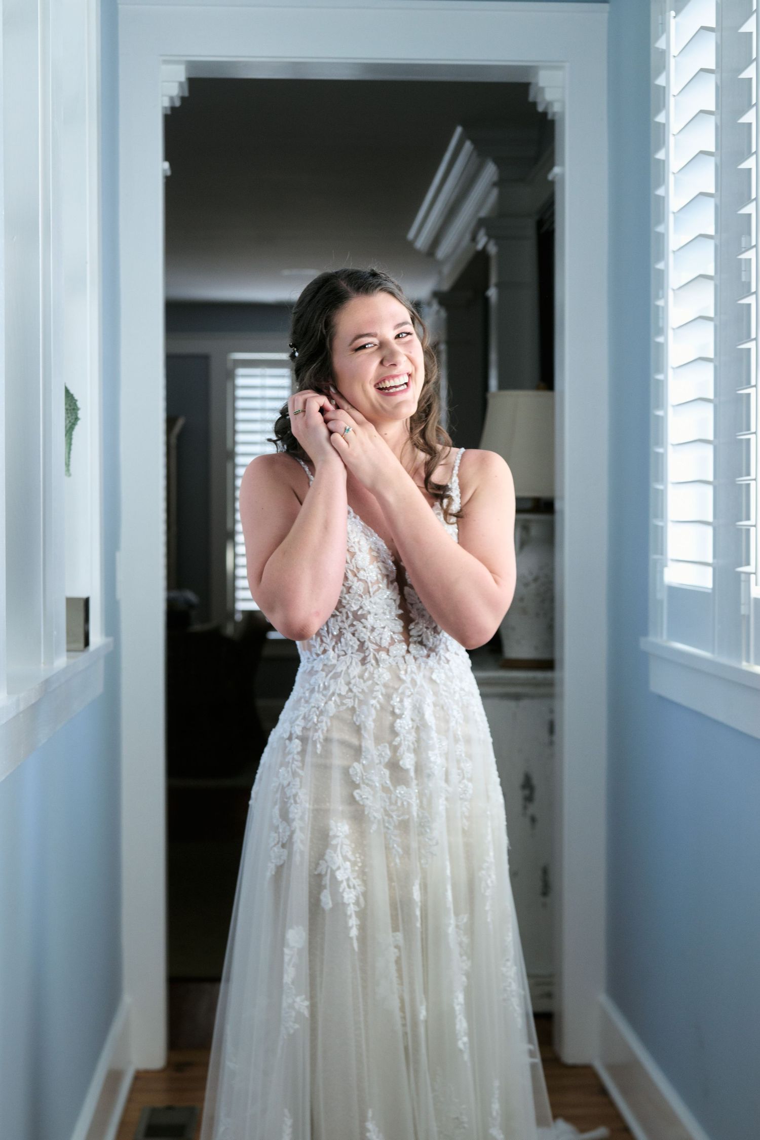 A bride wearing a white lace wedding gown stands in a sunlit hallway by shuttered windows.