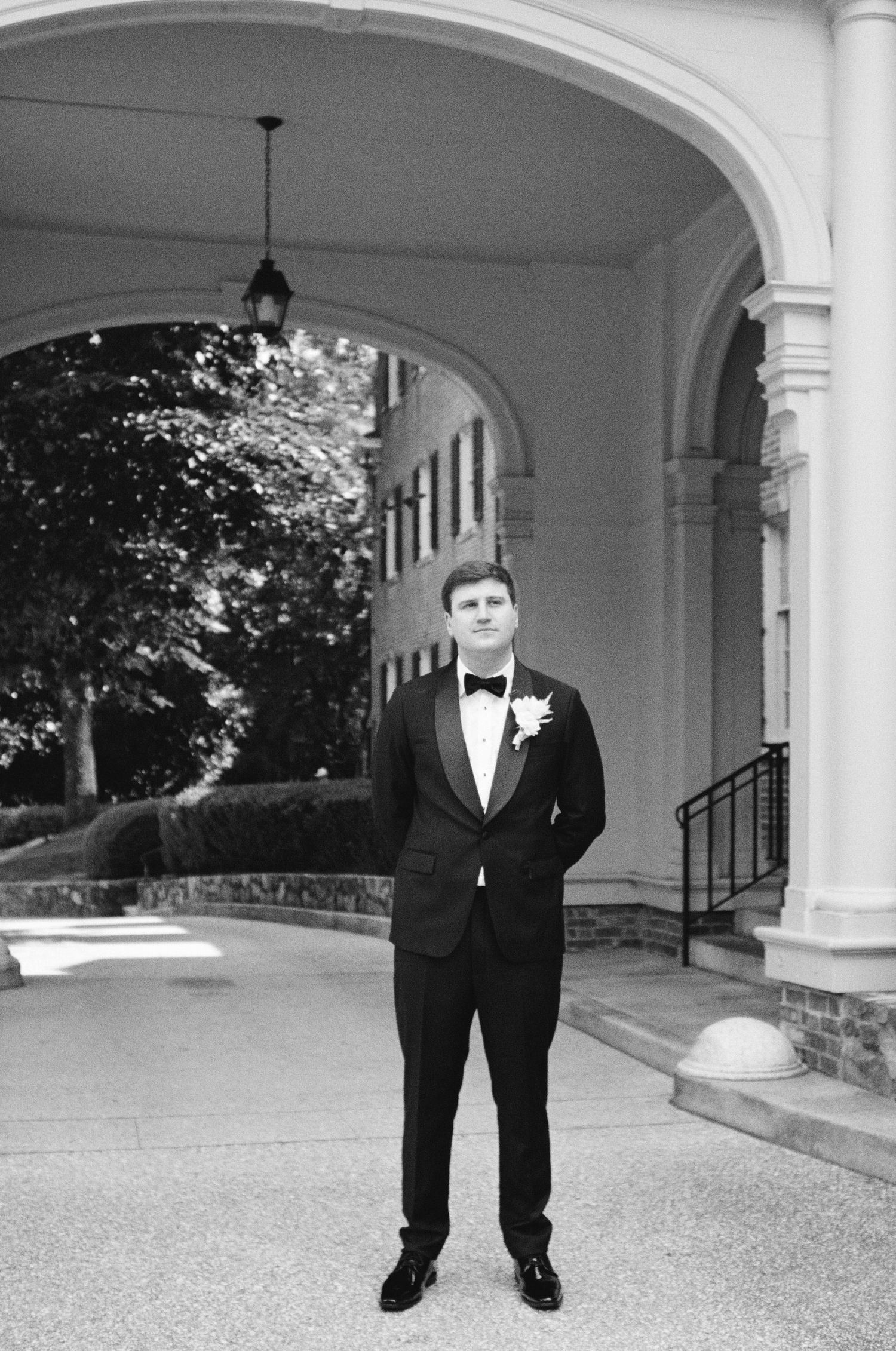 Classic black and white portrait of groom in tuxedo standing under historic columned archway.