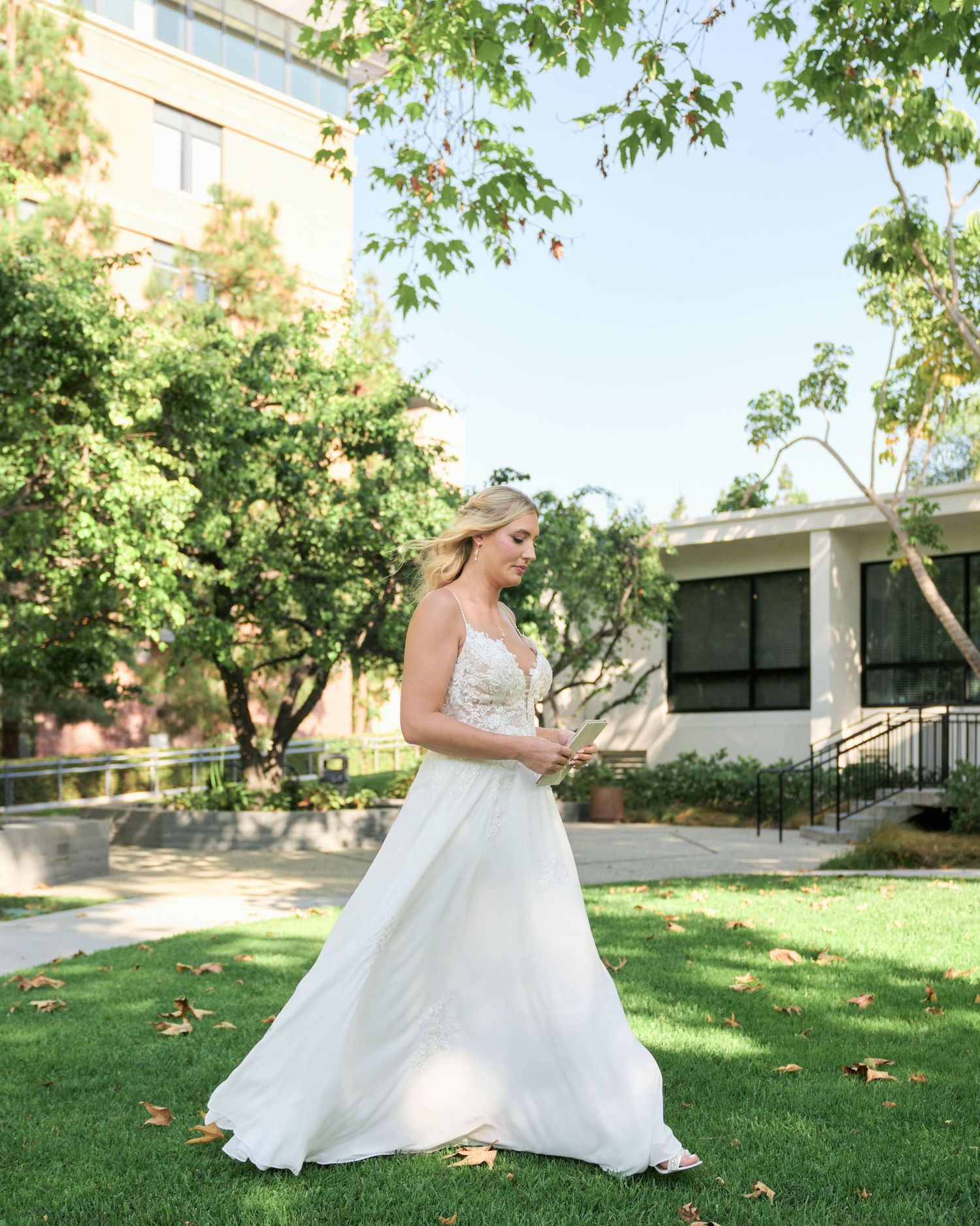 A bride in a flowing white dress poses outdoors on a sunny day with trees and a building in the background.