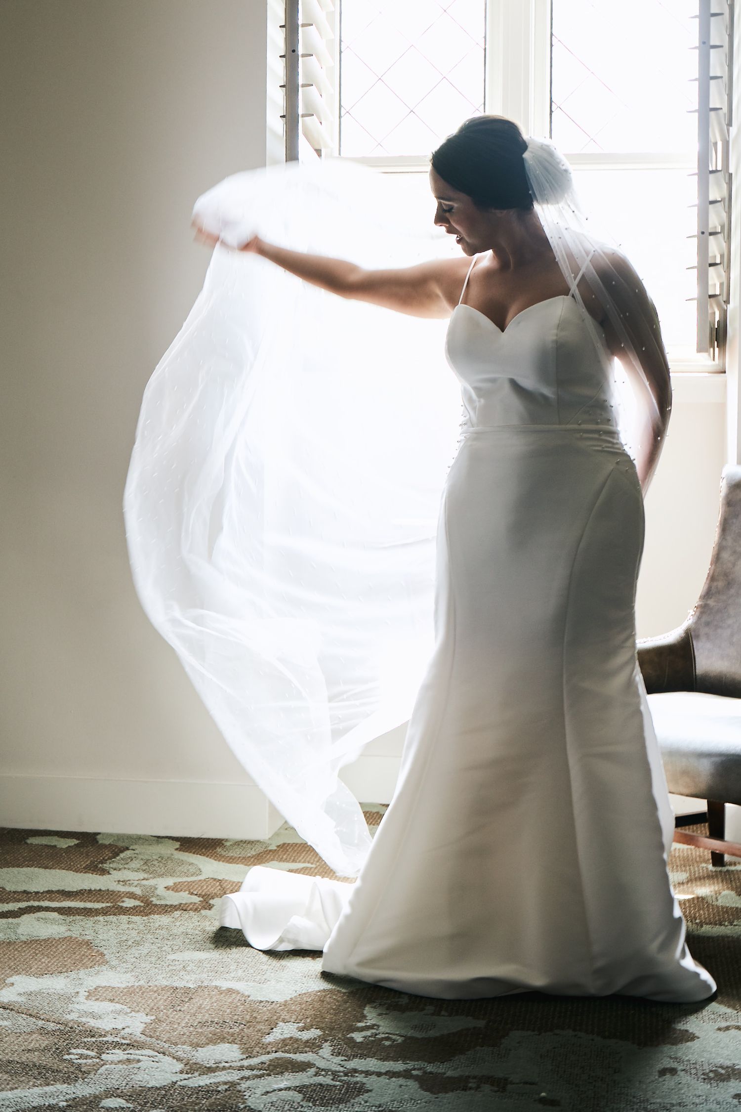 A bride in a white mermaid wedding dress adjusts her flowing veil by a window in a romantic pose.