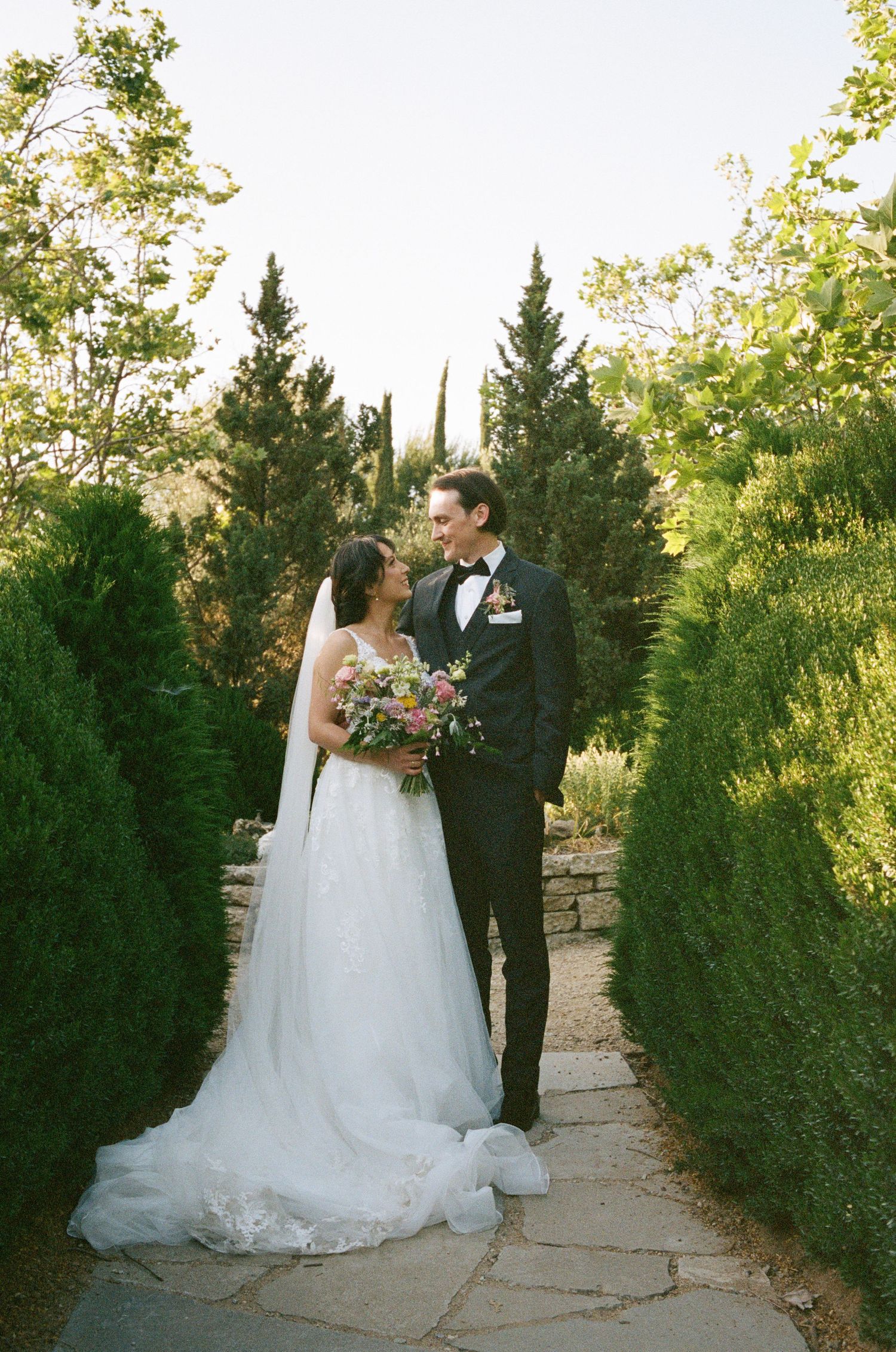 A couple in wedding attire stands on a garden path lined with evergreen hedges while holding a colorful bouquet.