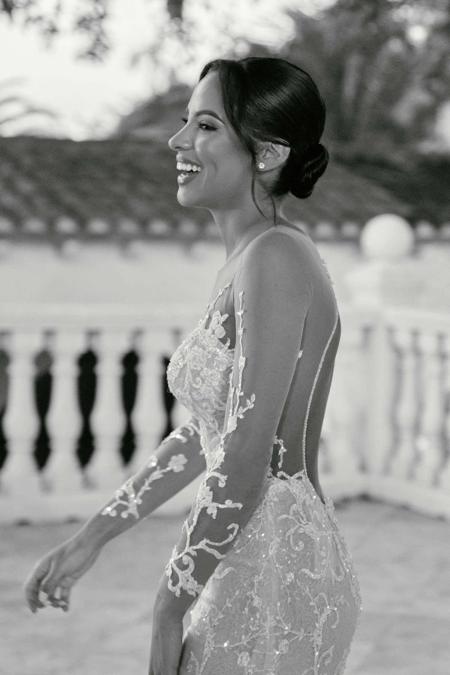 Black and white photo of a bride in a delicate lace gown smiling against an outdoor balustrade setting.