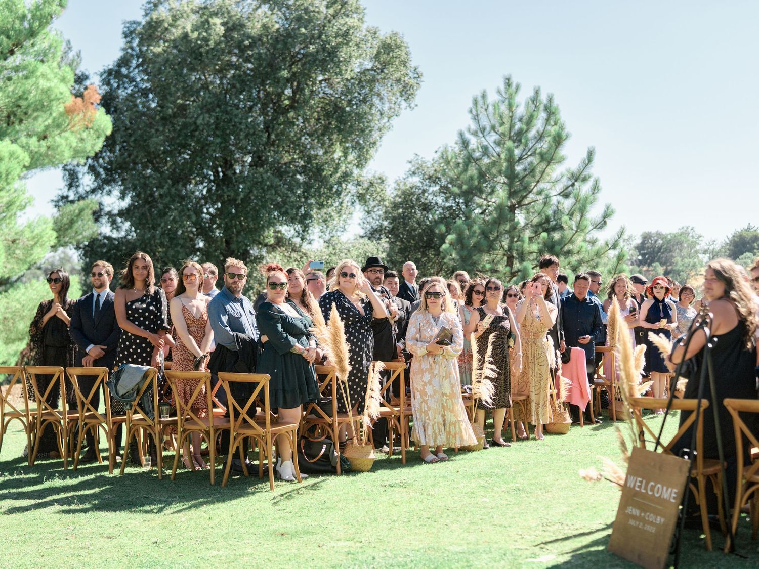 Wedding guests seated in wooden chairs at an outdoor ceremony under sunny skies.