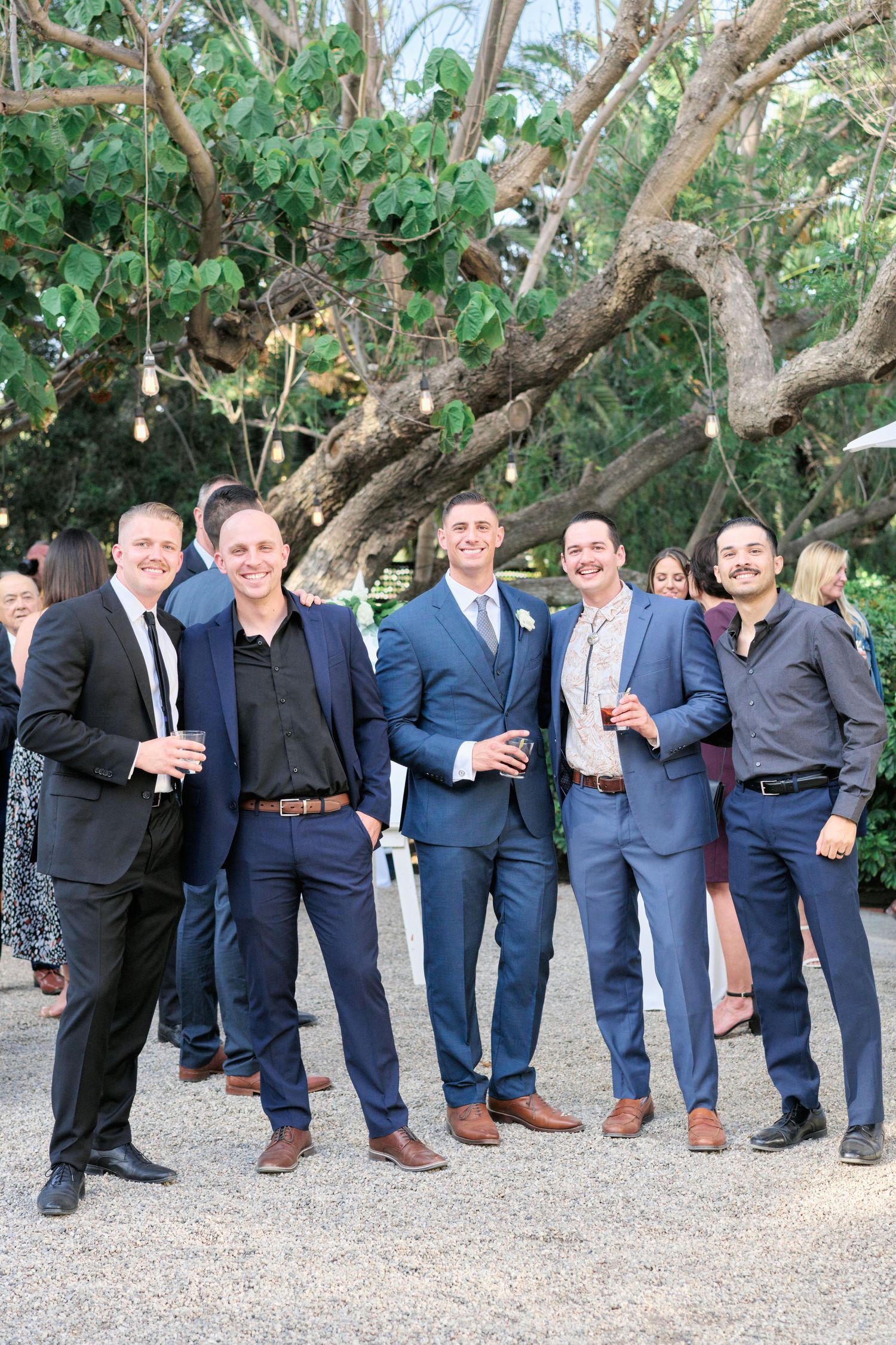 A group of men in formal attire pose together during a wedding celebration under tree branches.