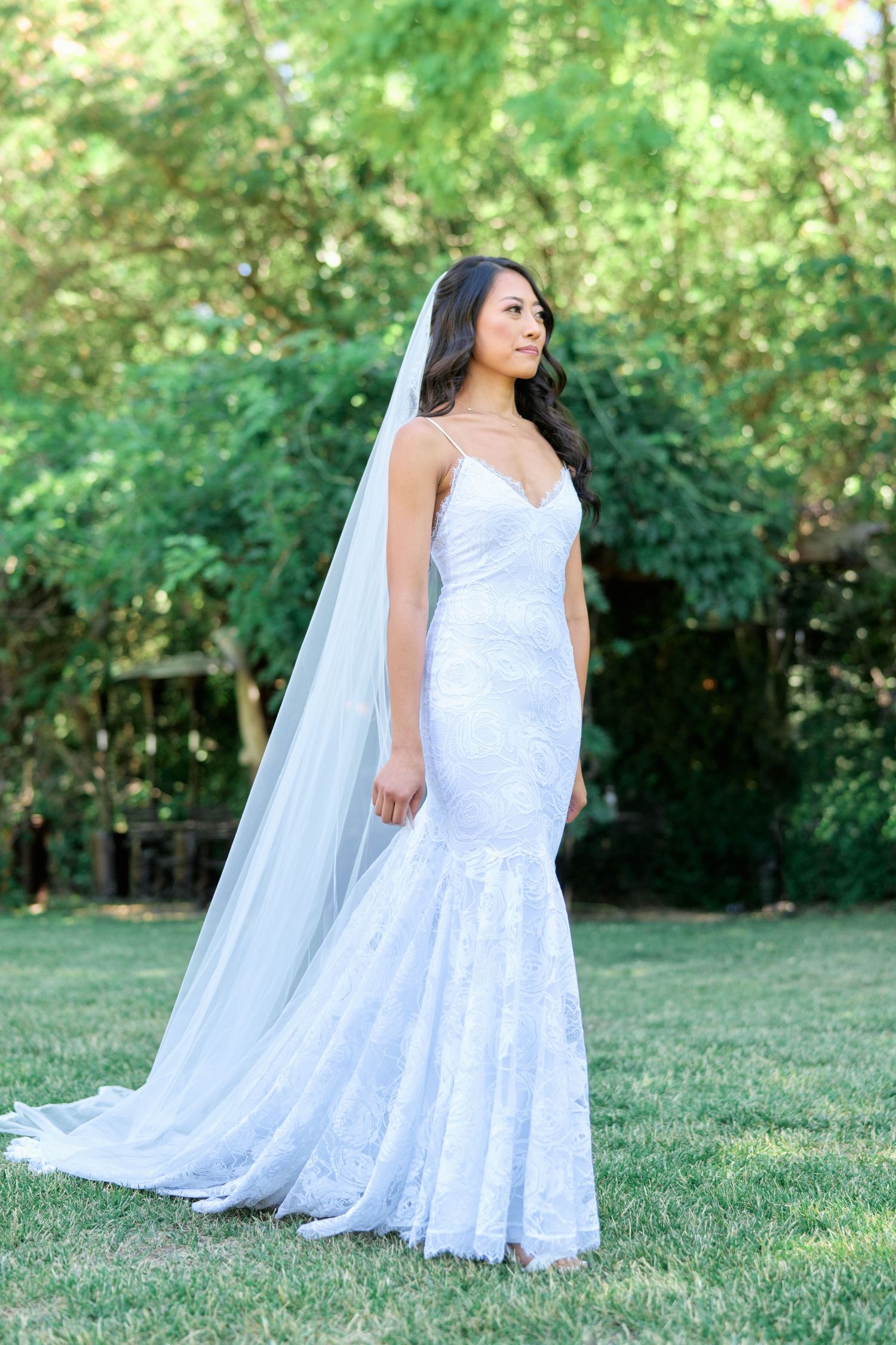 A bride in a white lace mermaid gown and veil poses in a garden setting.