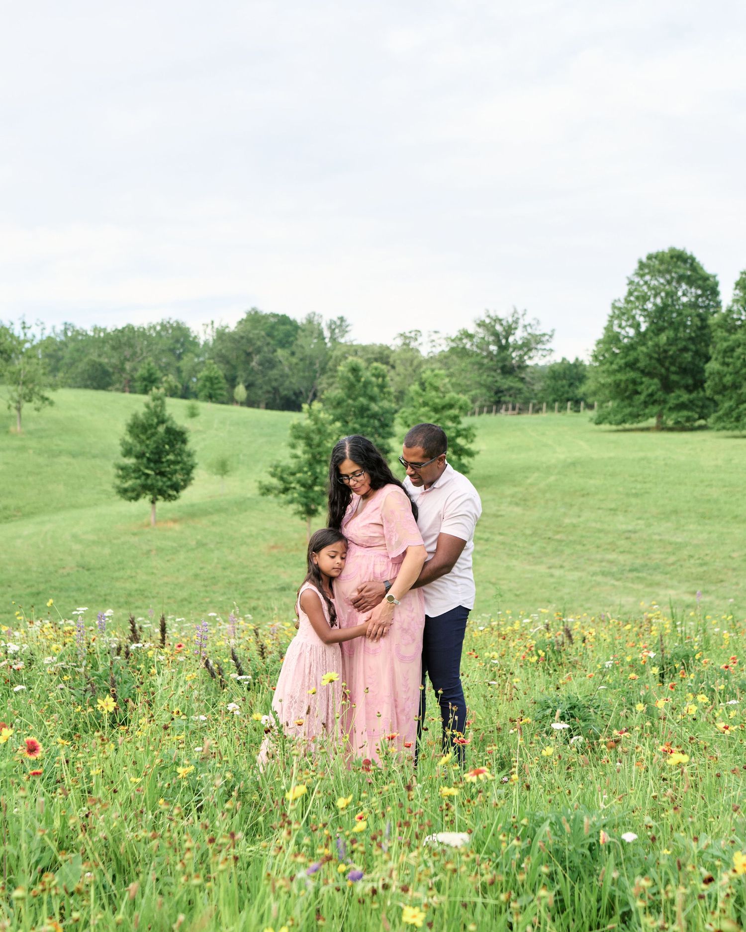 A pregnant woman in a pink dress stands with her partner and young daughter in a colorful wildflower meadow.