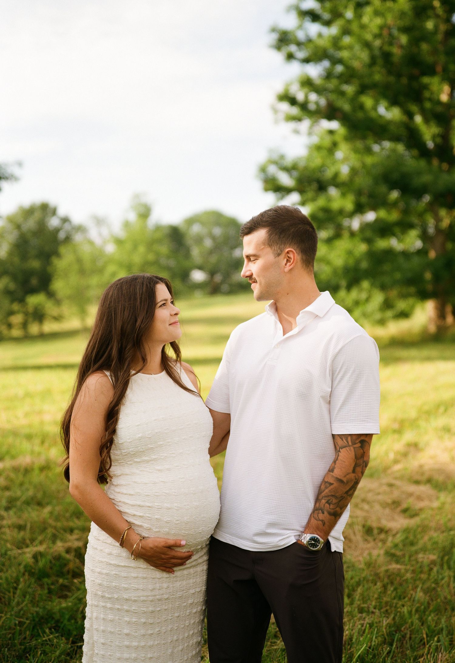 Pregnant couple in white outfits sharing a tender moment in a sunny green field, maternity photo session outdoors.