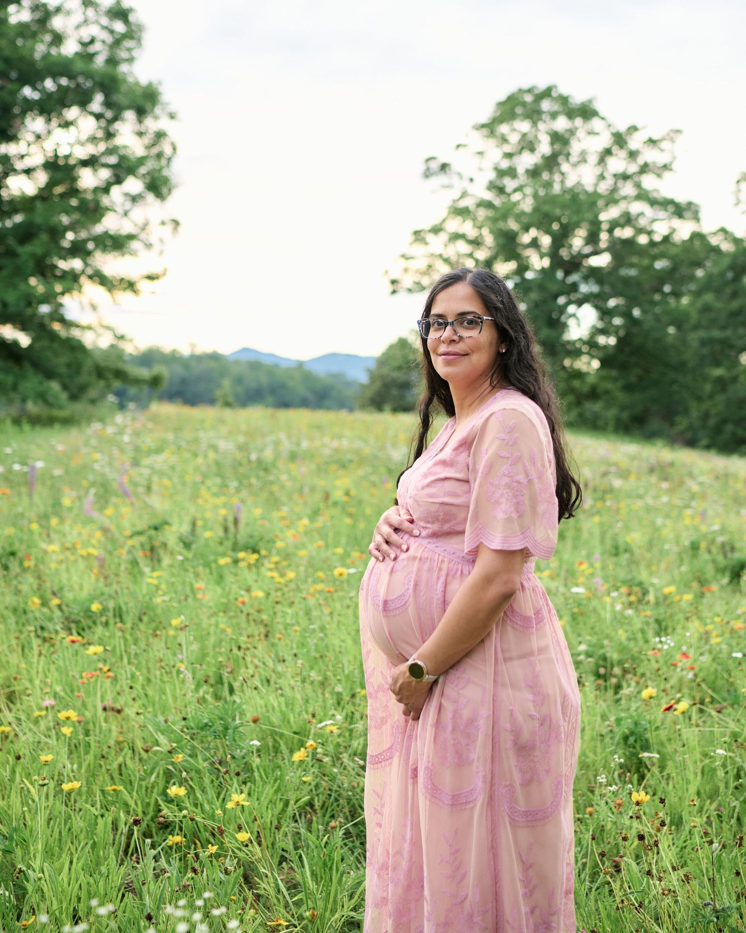 Pregnant woman in pink floral maxi dress standing in a wildflower meadow with mountains and trees in the background.