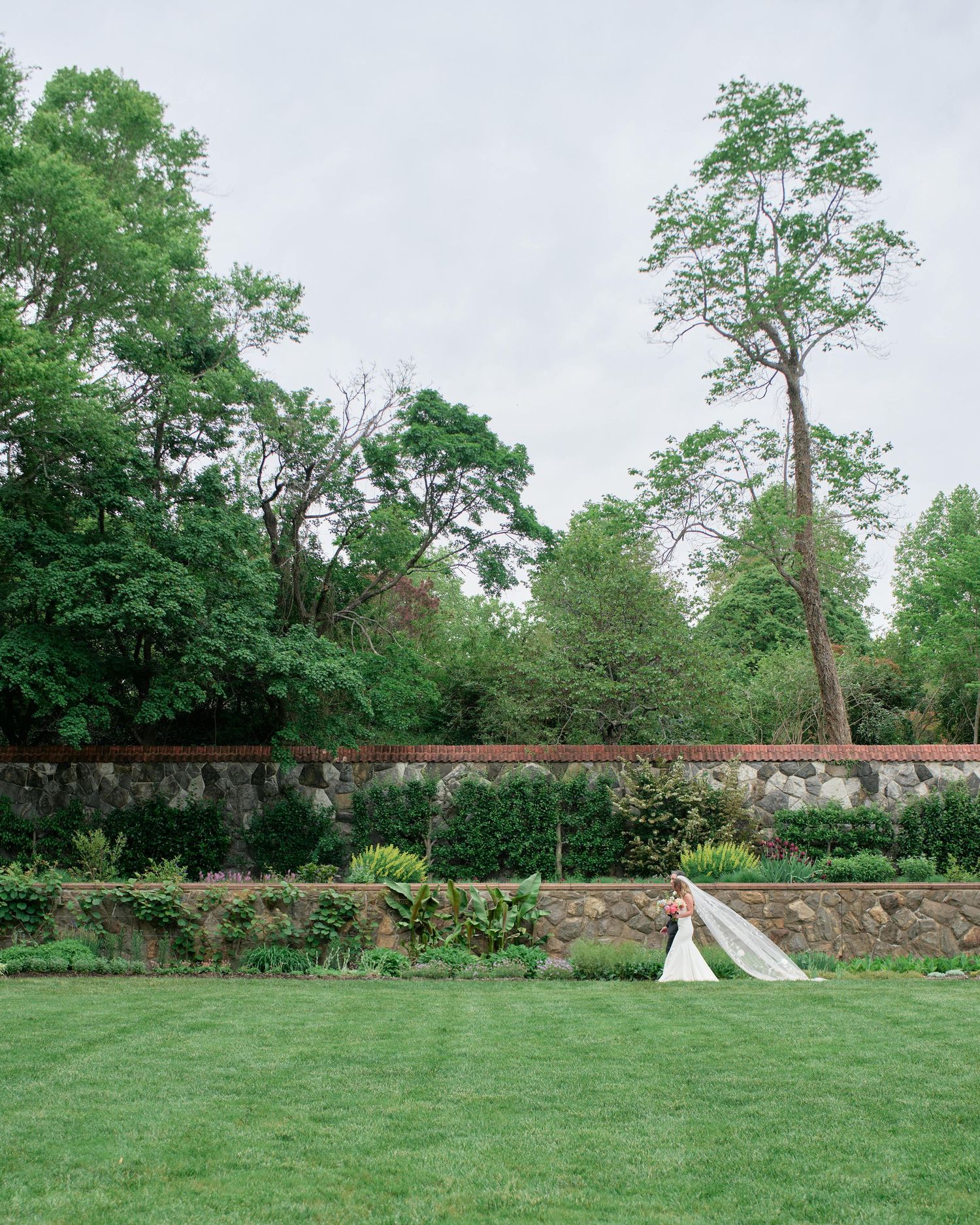 A panoramic view of a lush green lawn bordered by stone walls and tall trees with two small teepees in the distance.