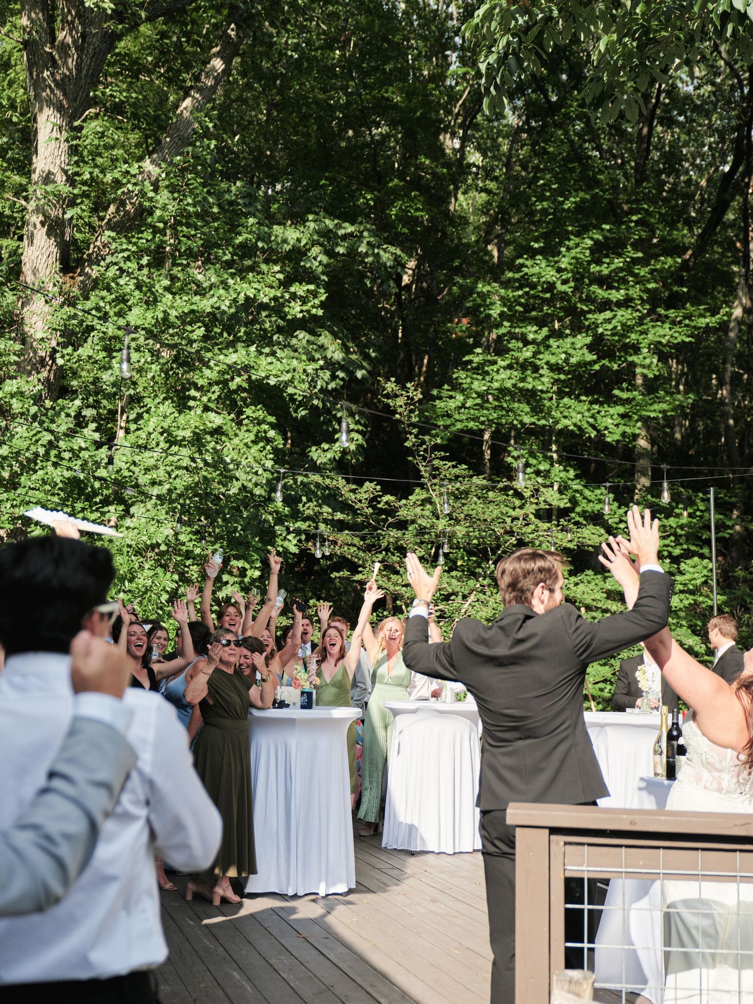 Guests raise hands in celebration at an outdoor wedding reception on a wooden deck surrounded by lush green trees.