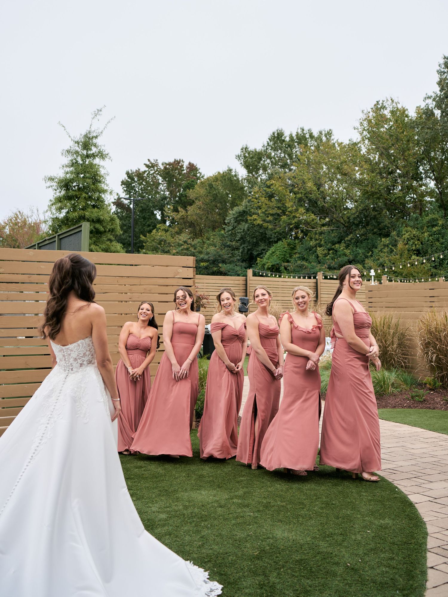 Bride in white gown reveals dress to bridesmaids in dusty rose gowns during outdoor first look ceremony.