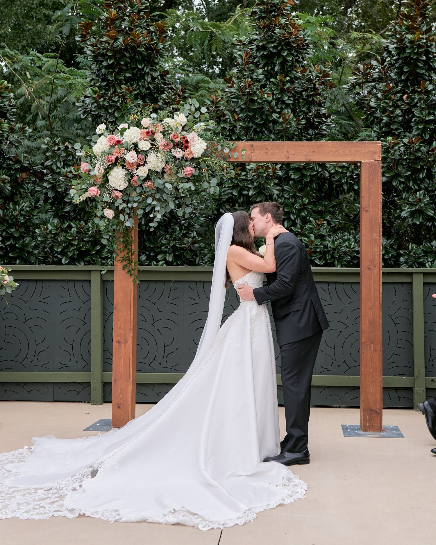 Bride and groom share first kiss under wooden arch with pink floral arrangement at outdoor wedding ceremony.