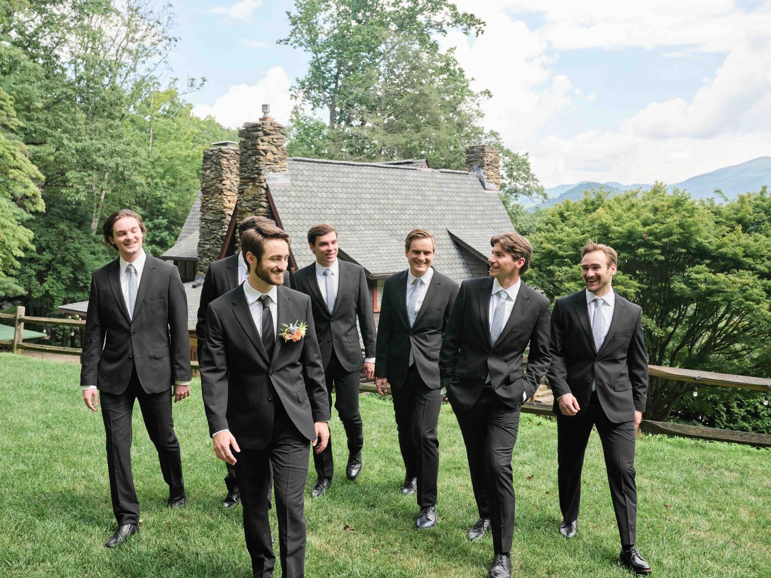 Groom and groomsmen in dark suits walking outdoors near a stone chapel surrounded by lush green trees.