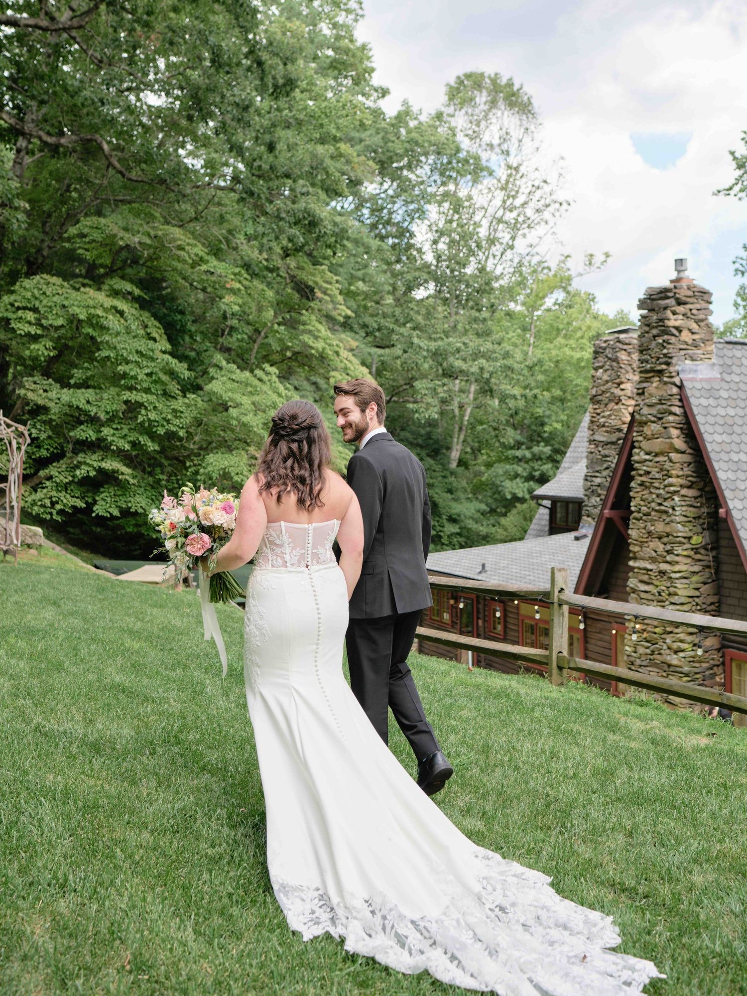 Bride in lace gown with bouquet and groom in gray suit walk toward rustic stone chapel surrounded by lush green trees.