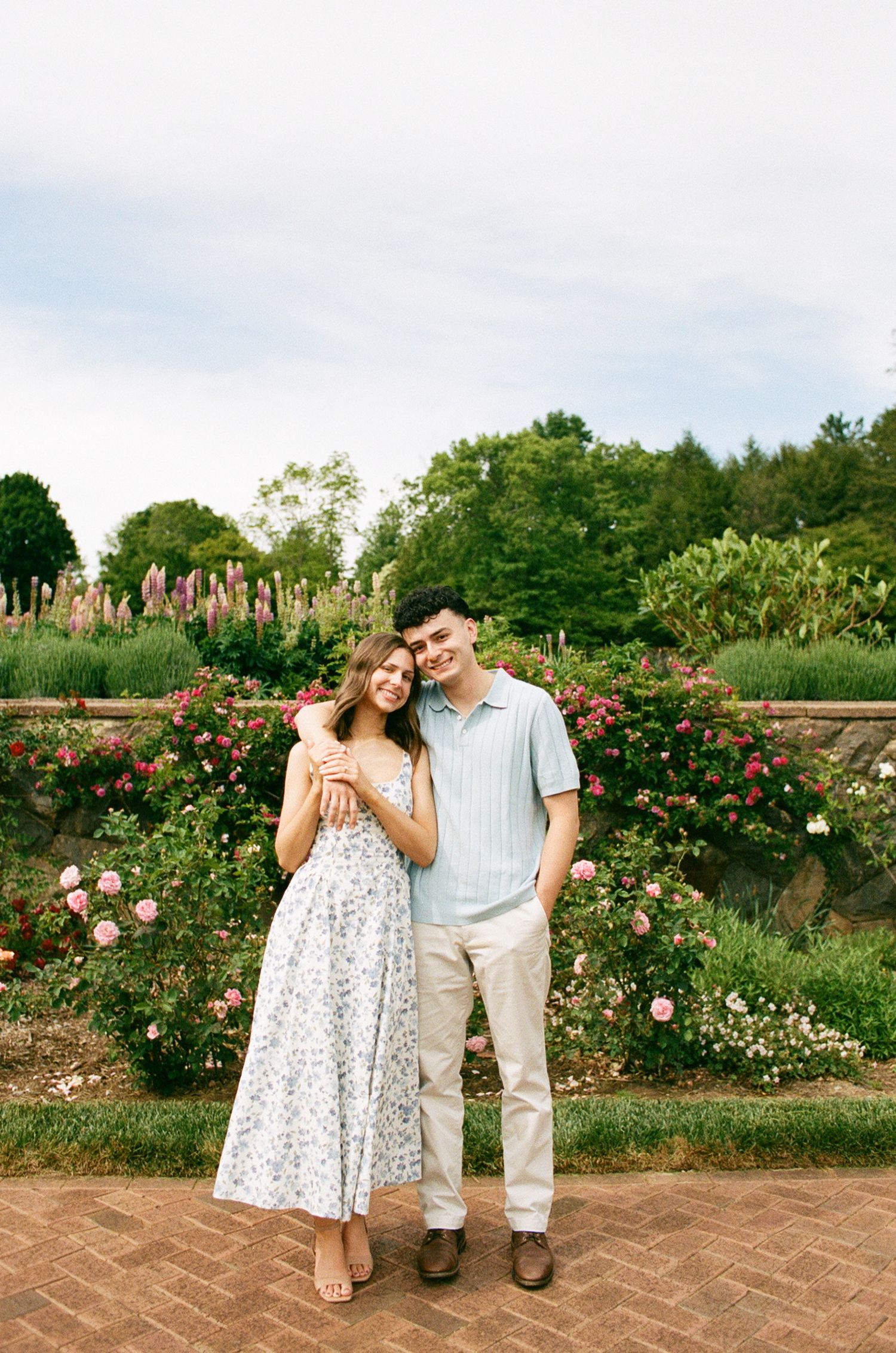 A couple poses together in a lush garden with blooming roses, the woman in a floral dress and man in a blue polo.