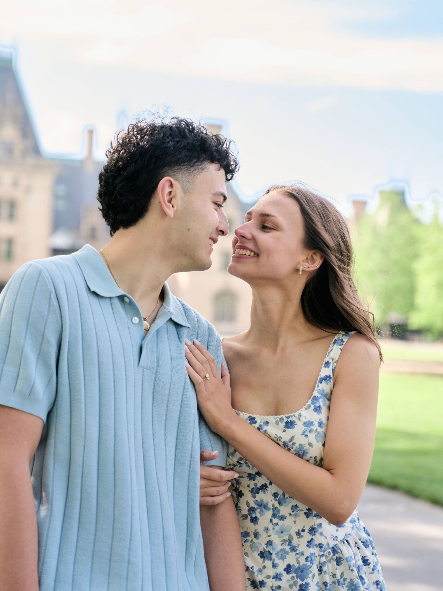 A smiling couple in a garden, woman in floral dress touches man's chest as they lean close together.