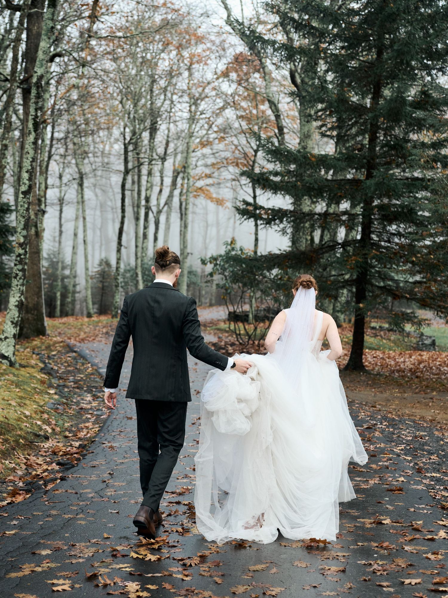 Newlyweds walking down autumn forest path holding dress train.