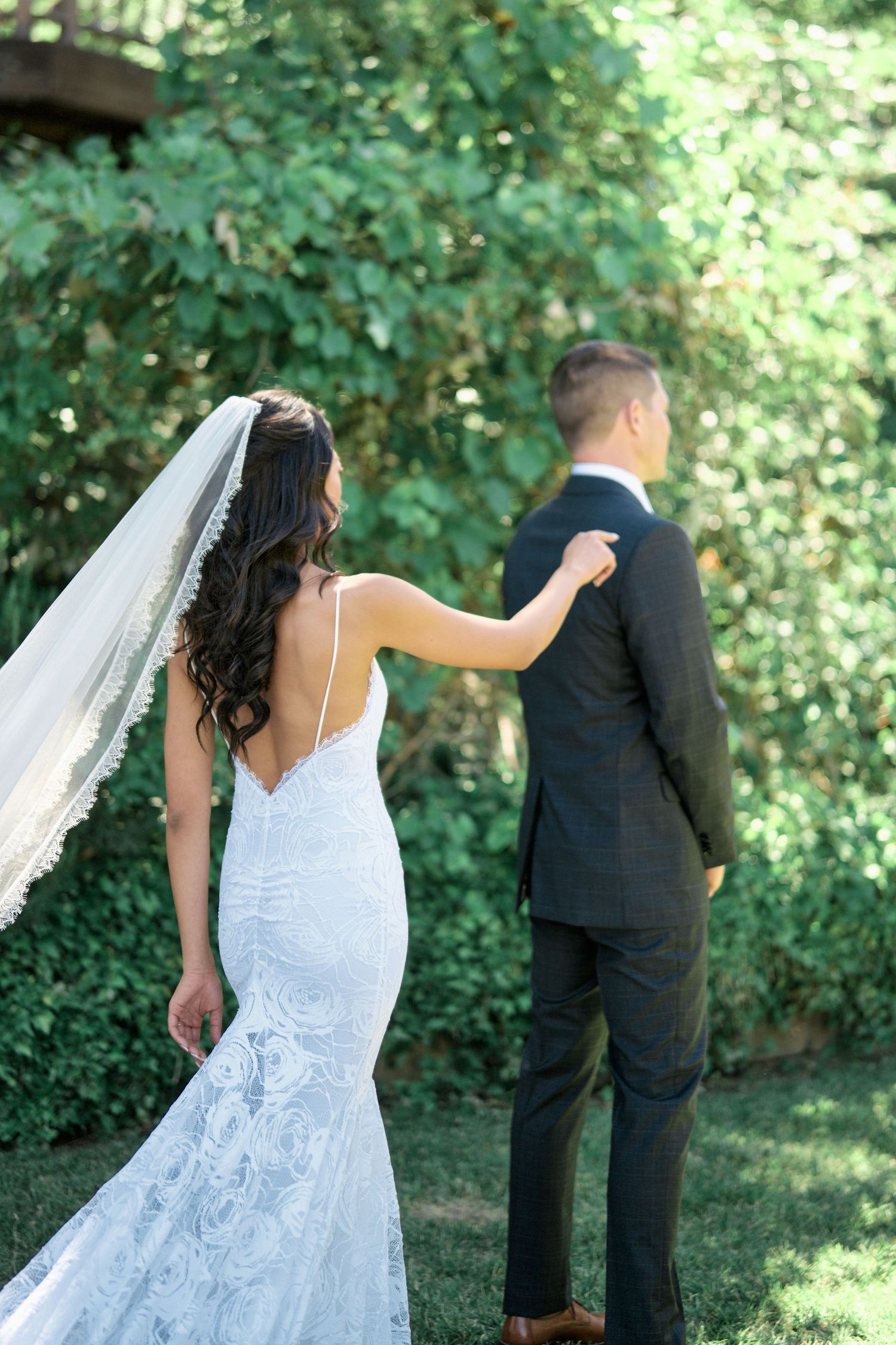 Bride in fitted lace gown touches groom's shoulder during first look in garden setting.