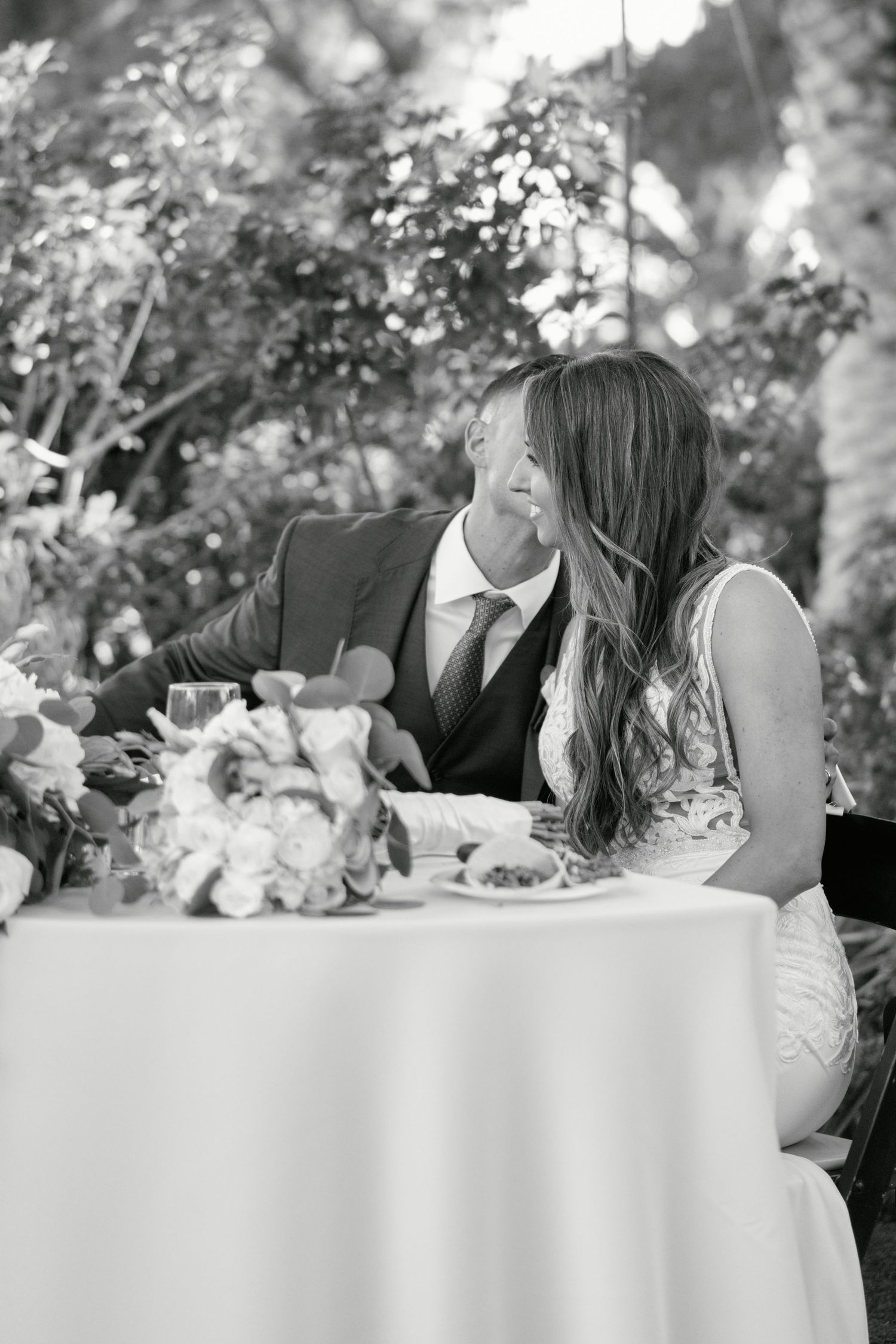 Black and white candid moment of a couple sharing a laugh at their reception table.