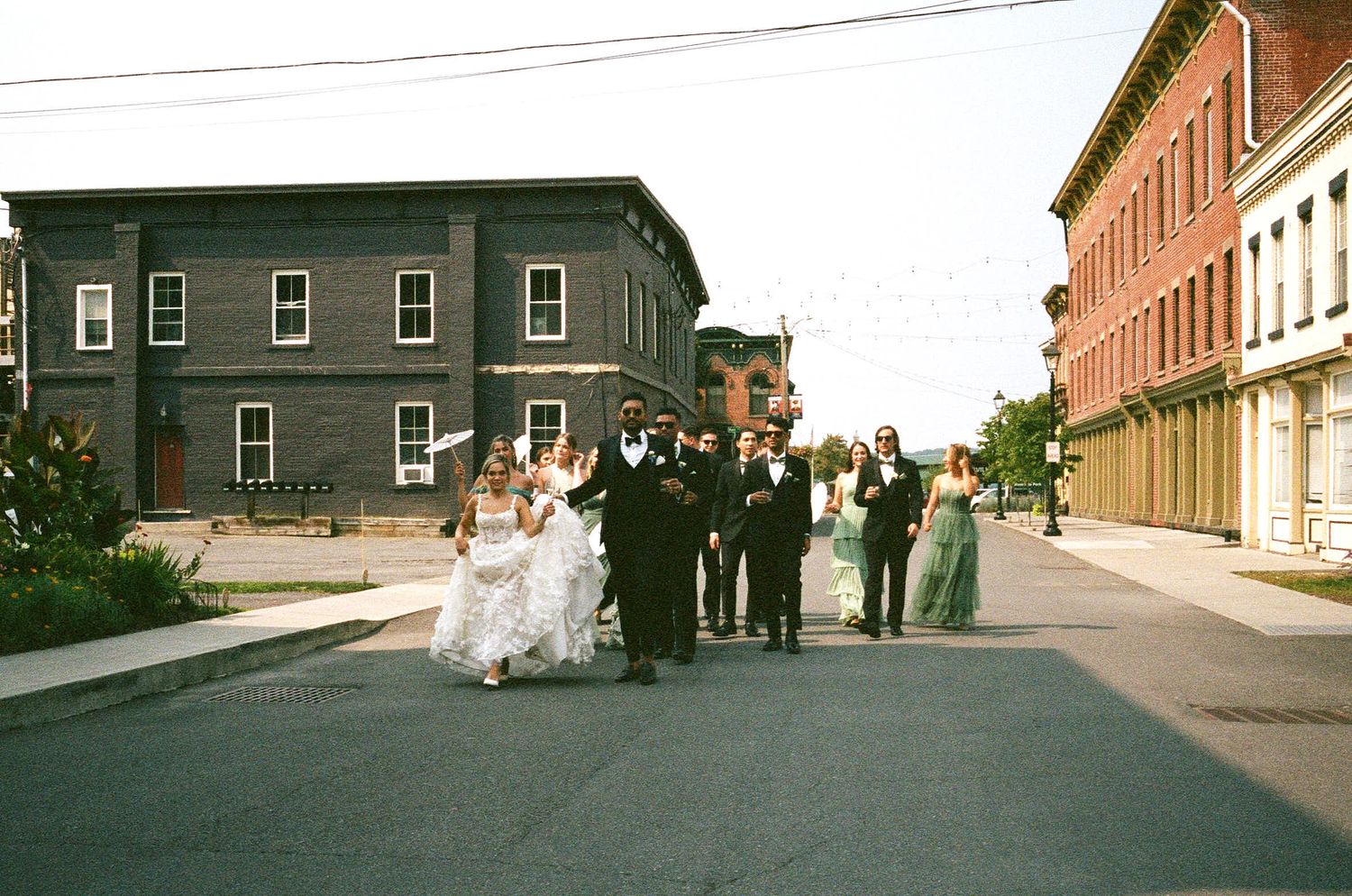 A wedding party gathered on a city street between historic brick and wooden buildings on a sunny day.