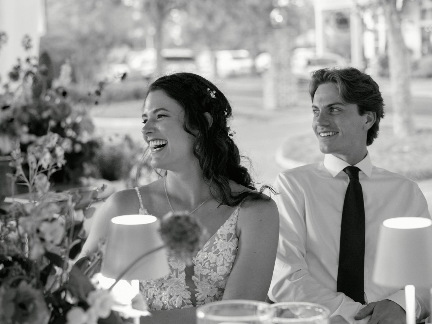 A black and white photo of a couple sharing a joyful moment at a wedding reception table with floral decorations.