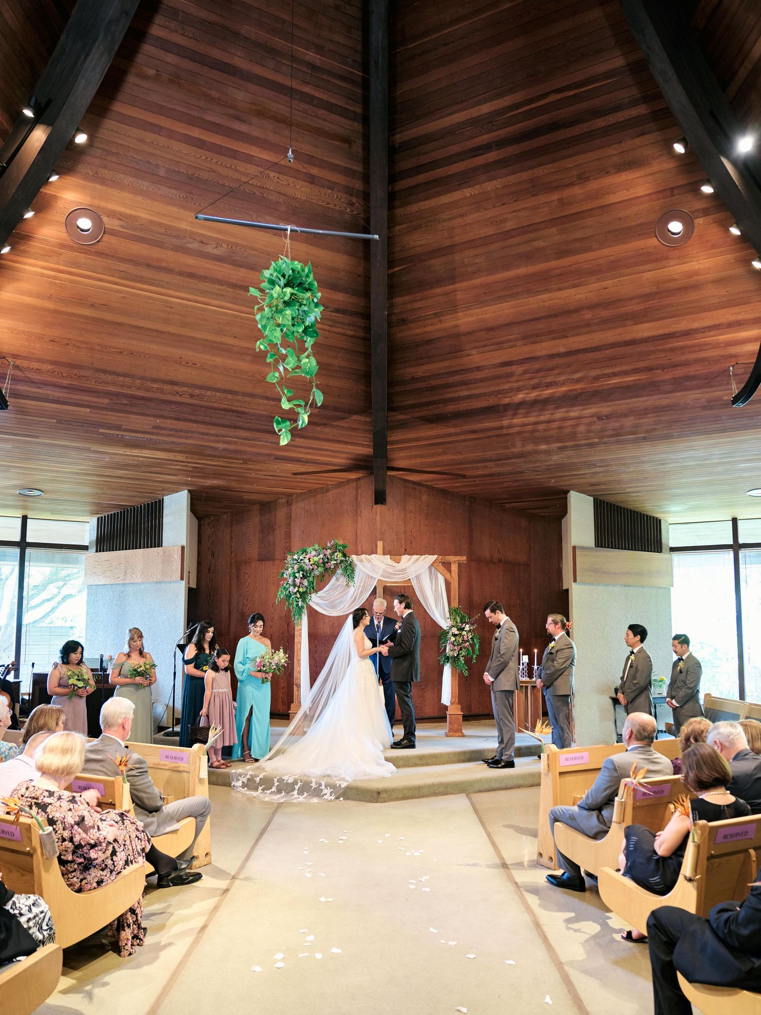 A wedding ceremony in progress inside a modern wooden chapel with hanging greenery.