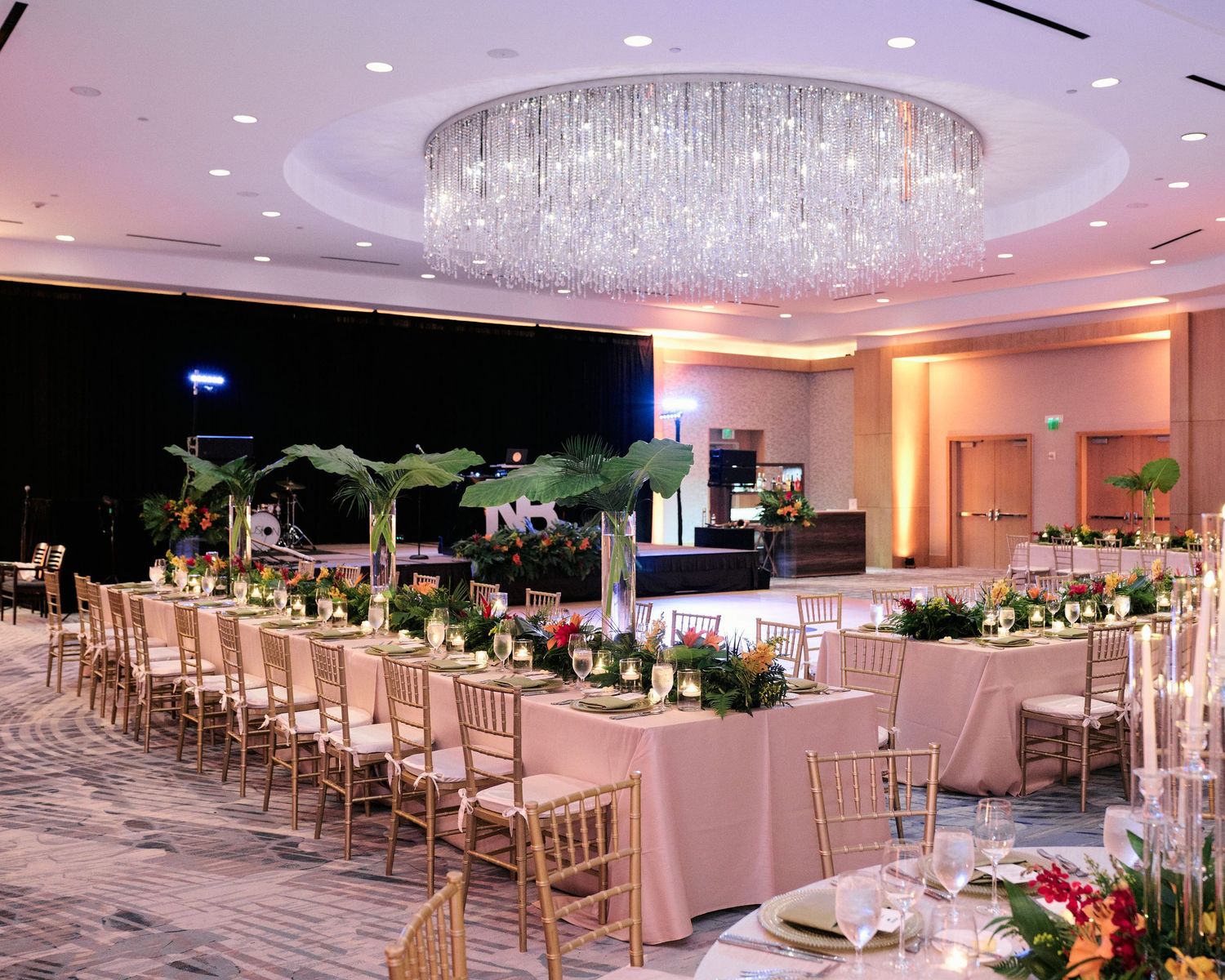 An elegant wedding reception setup with long tables, gold chiavari chairs, and a grand crystal chandelier overhead at the Ritz Carlton.