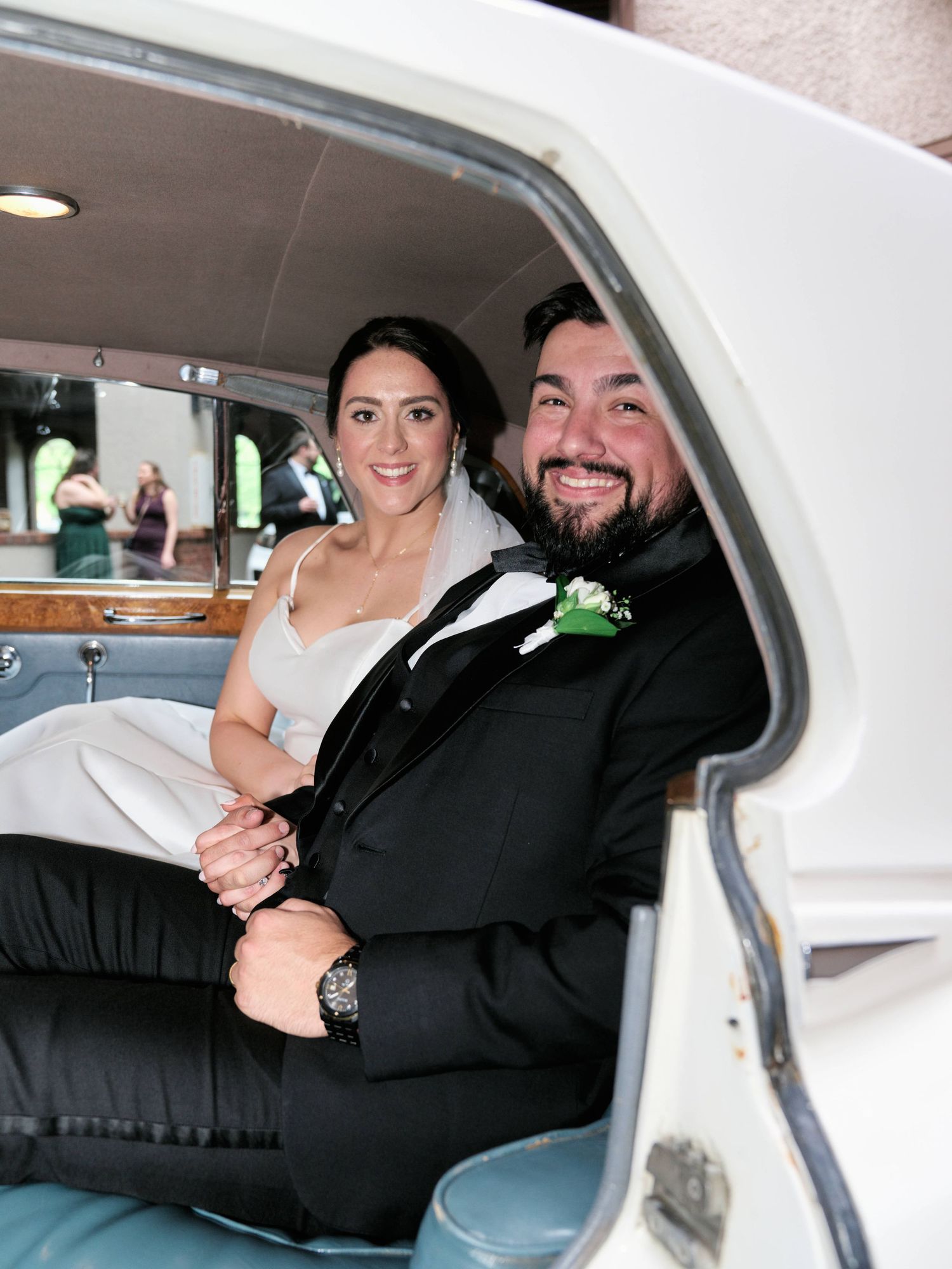 A happy couple sits together in the back seat of a white car during a wedding celebration.