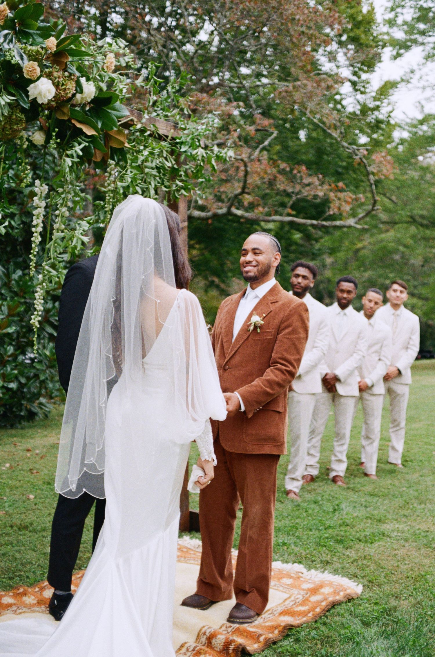 Outdoor wedding ceremony with bride in white dress and groom in brown suit standing under floral arch.