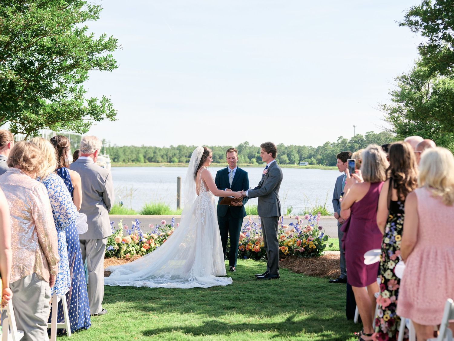 An outdoor lakeside wedding ceremony with guests gathered around floral arrangements and a scenic water view.