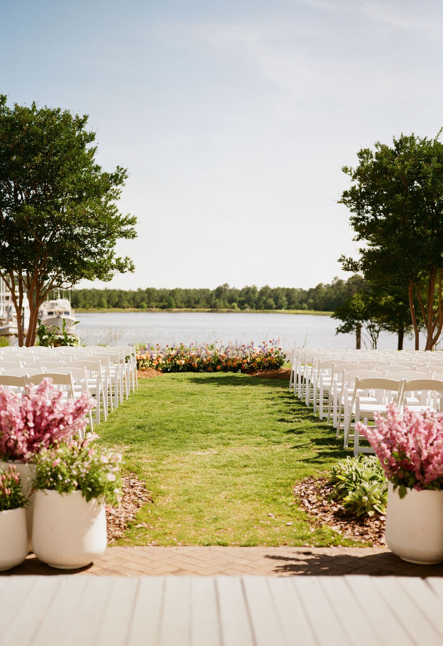 Lakefront wedding ceremony setup with white chairs and pink floral arrangements bordering a grassy aisle leading to the water.