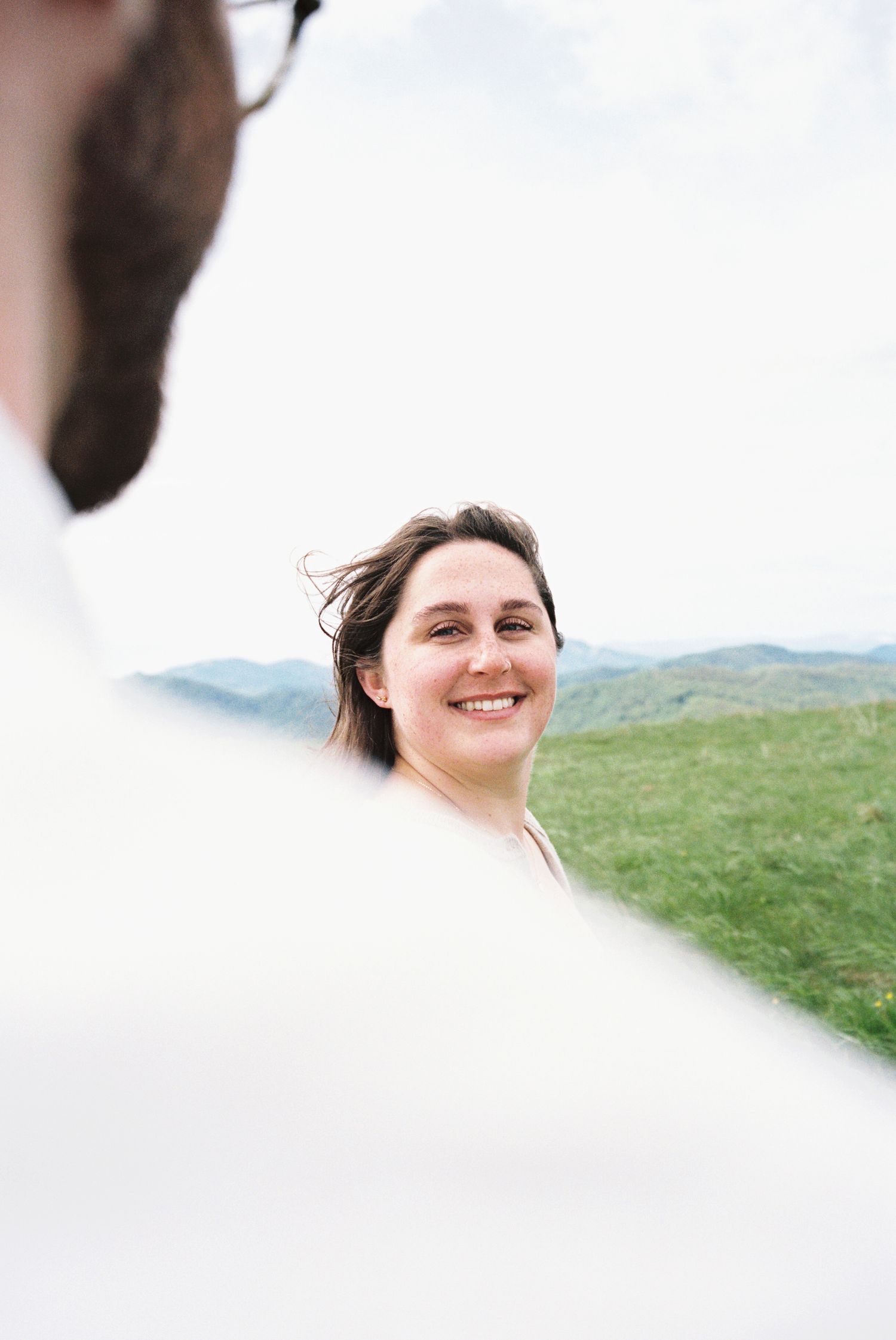 Two people sharing a moment outdoors on a grassy hillside with mountains visible in the background on a sunny day.