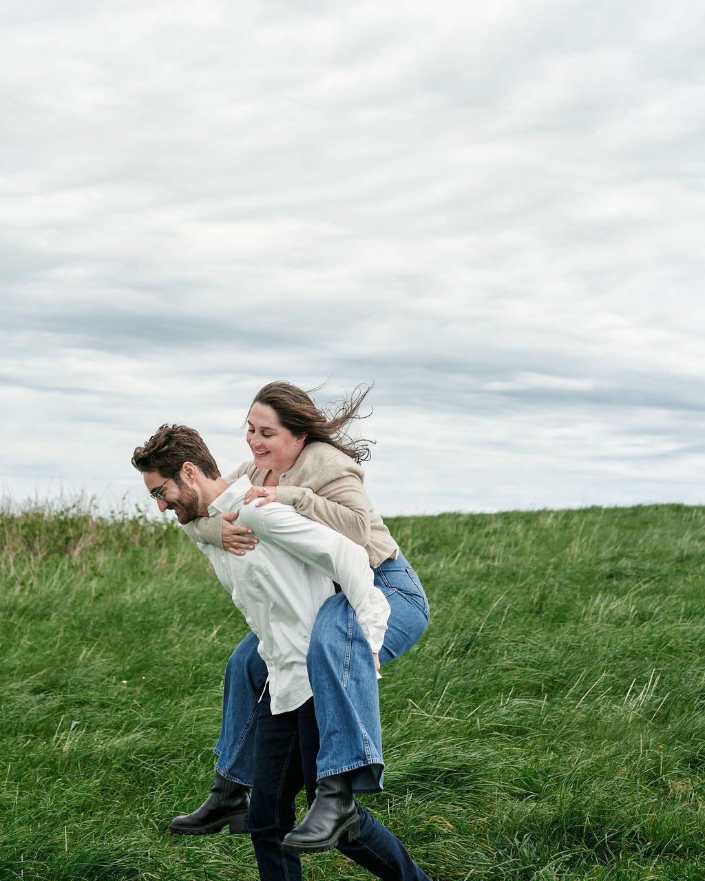 A couple enjoys a playful piggyback ride in a windswept green field under cloudy skies.