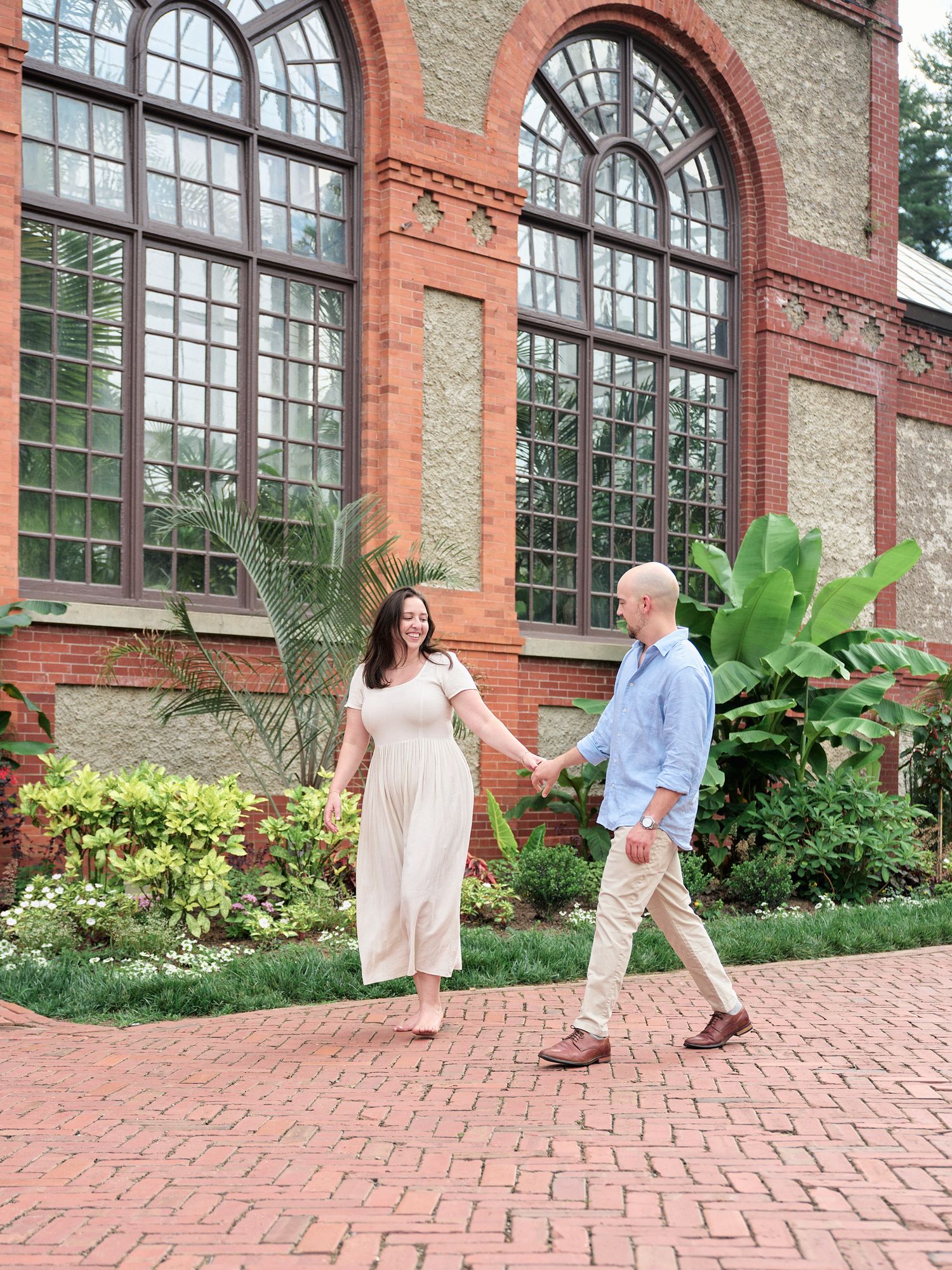 A couple strolls along a brick pathway in front of a historic brick building with arched windows and lush gardens.