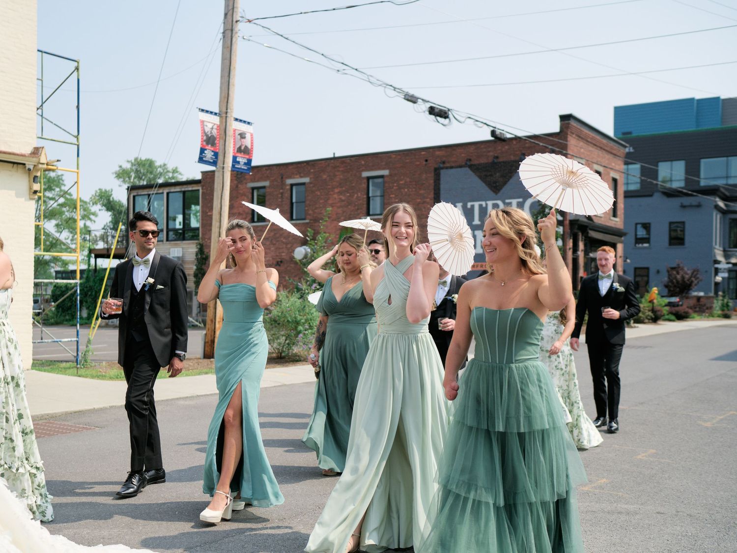 A wedding party walks down a street in sage green dresses and formal attire during a sunny day.