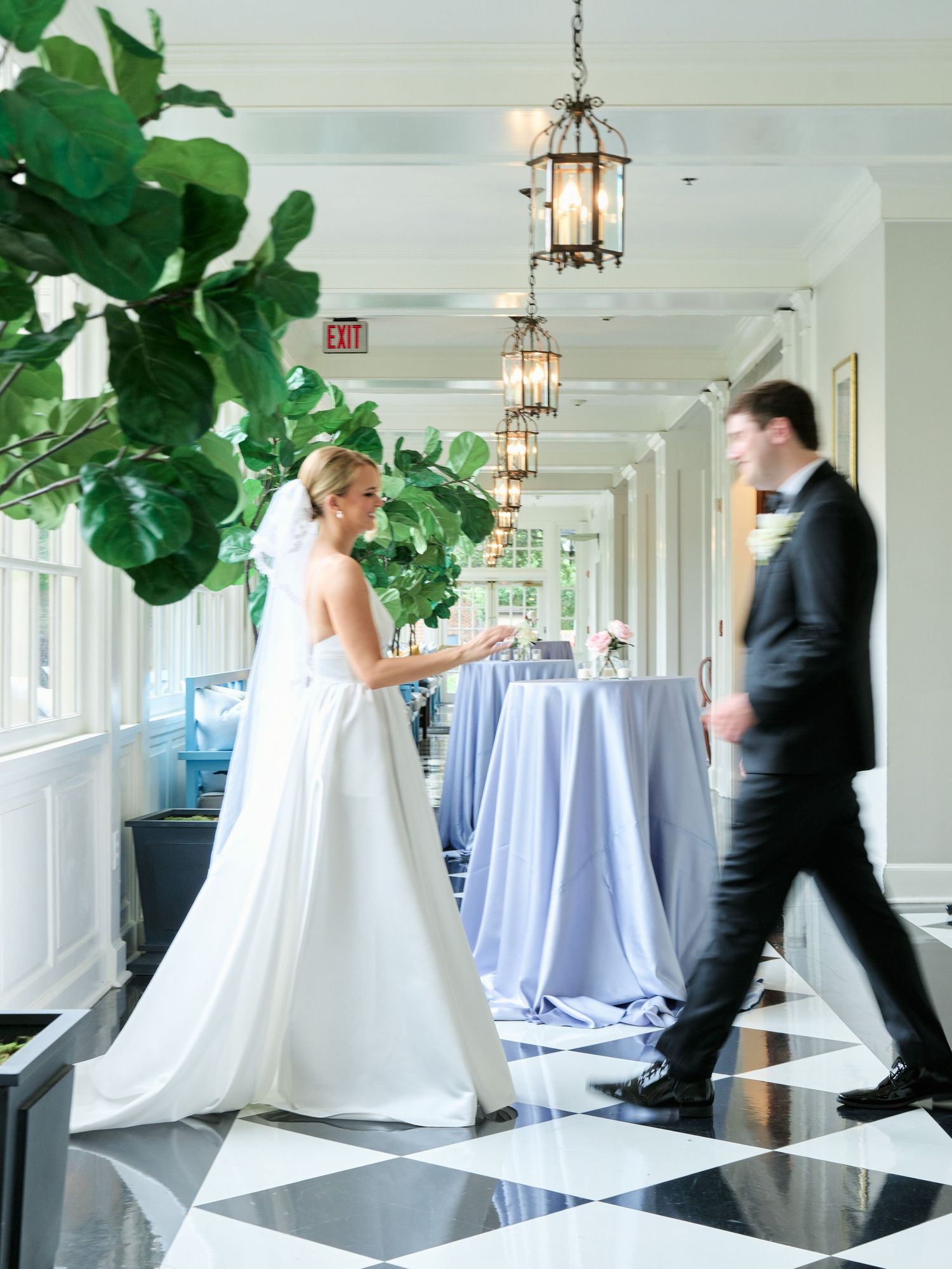Elegant wedding venue hallway with checkered floors, hanging lanterns and large fiddle leaf fig plants.