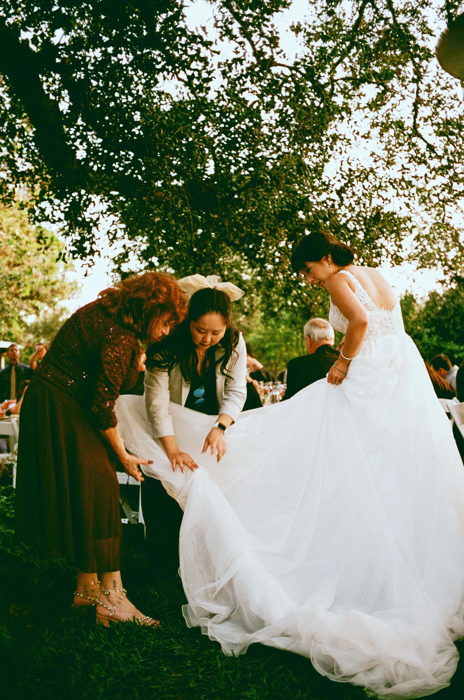 Wedding guests help arrange the bride's flowing white dress outdoors under a tree.