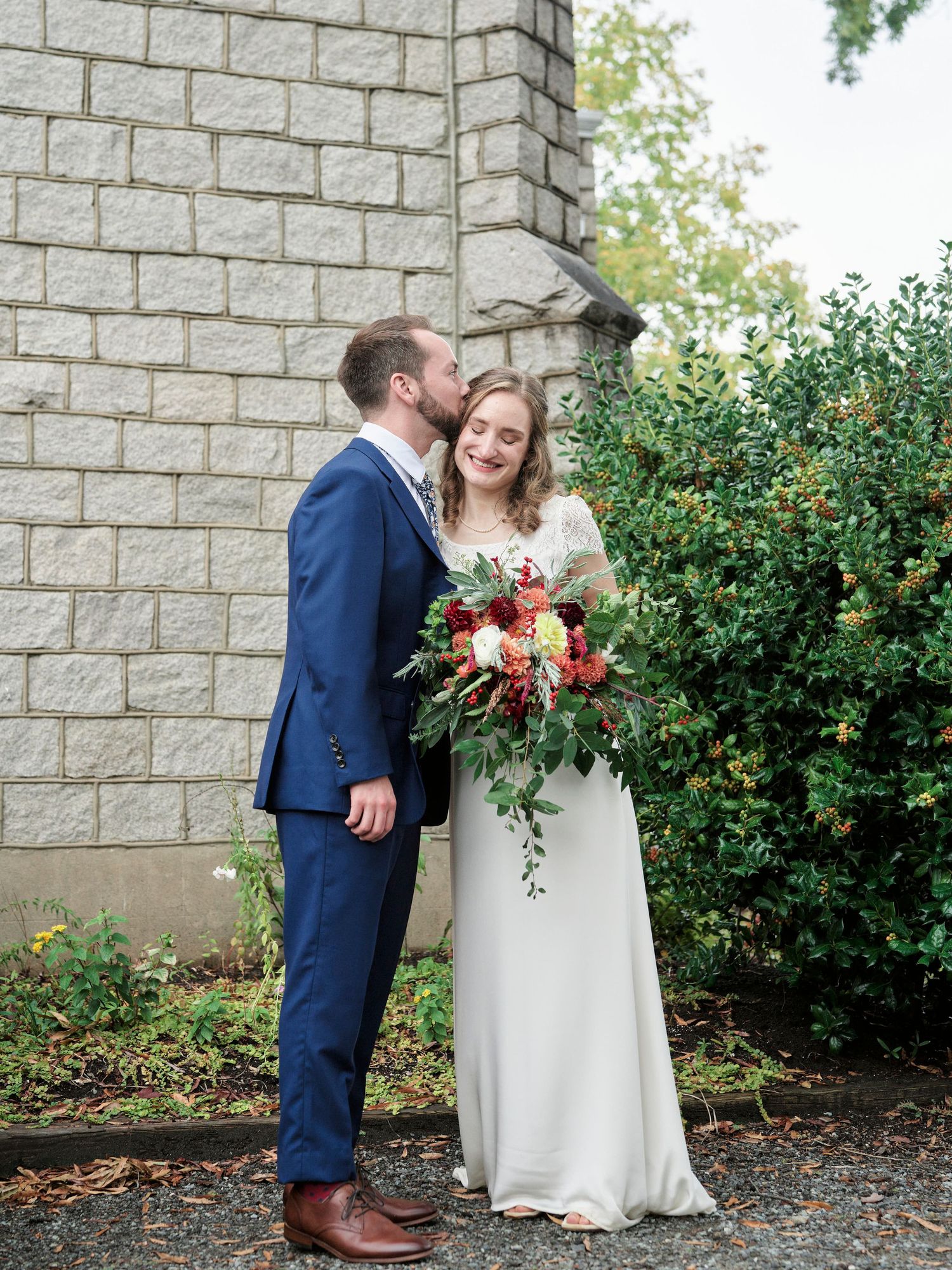 A couple shares an intimate moment outside a stone church with a beautiful floral bouquet.