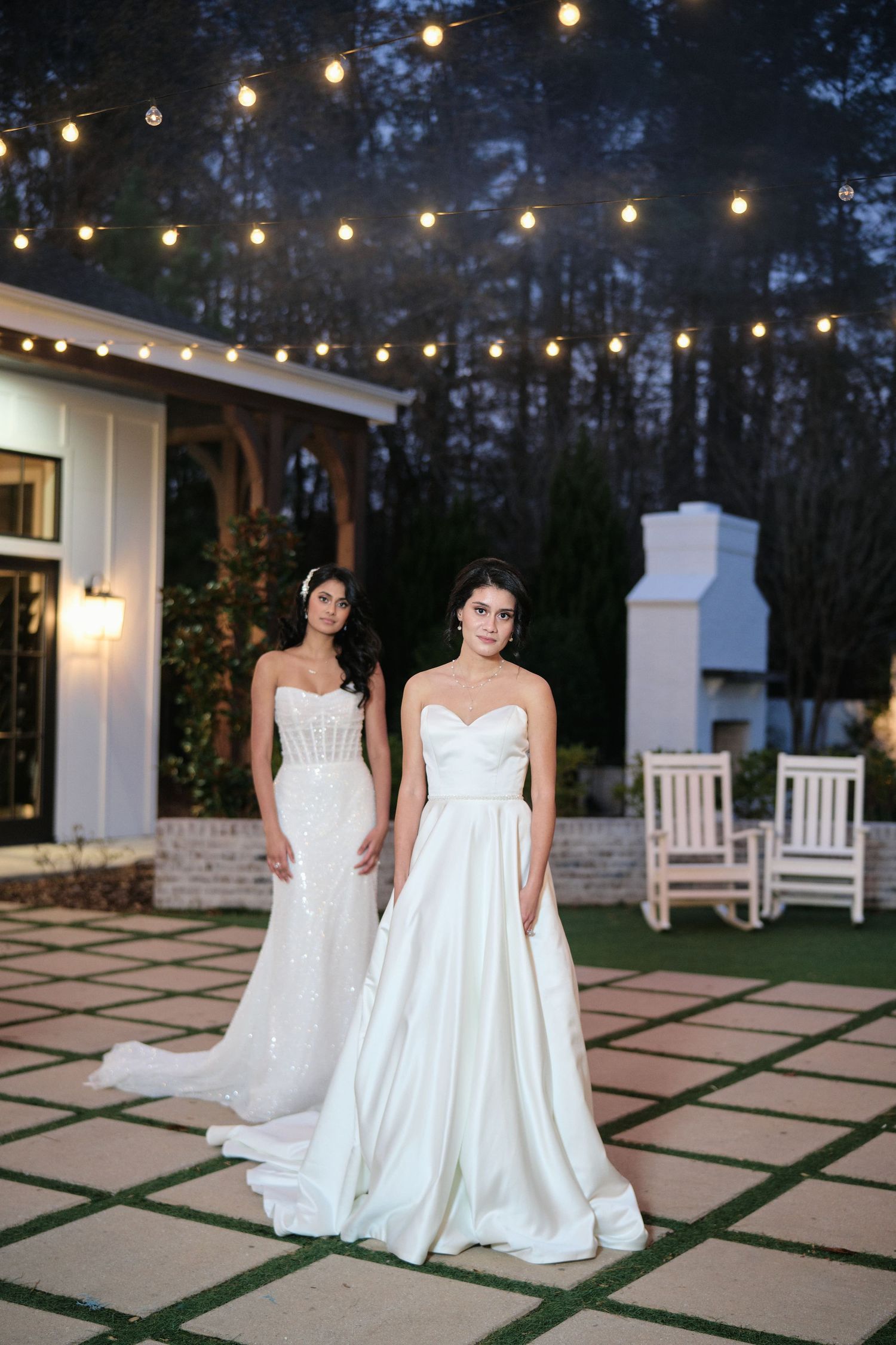 Two brides in elegant white gowns pose on a patio under string lights at dusk.