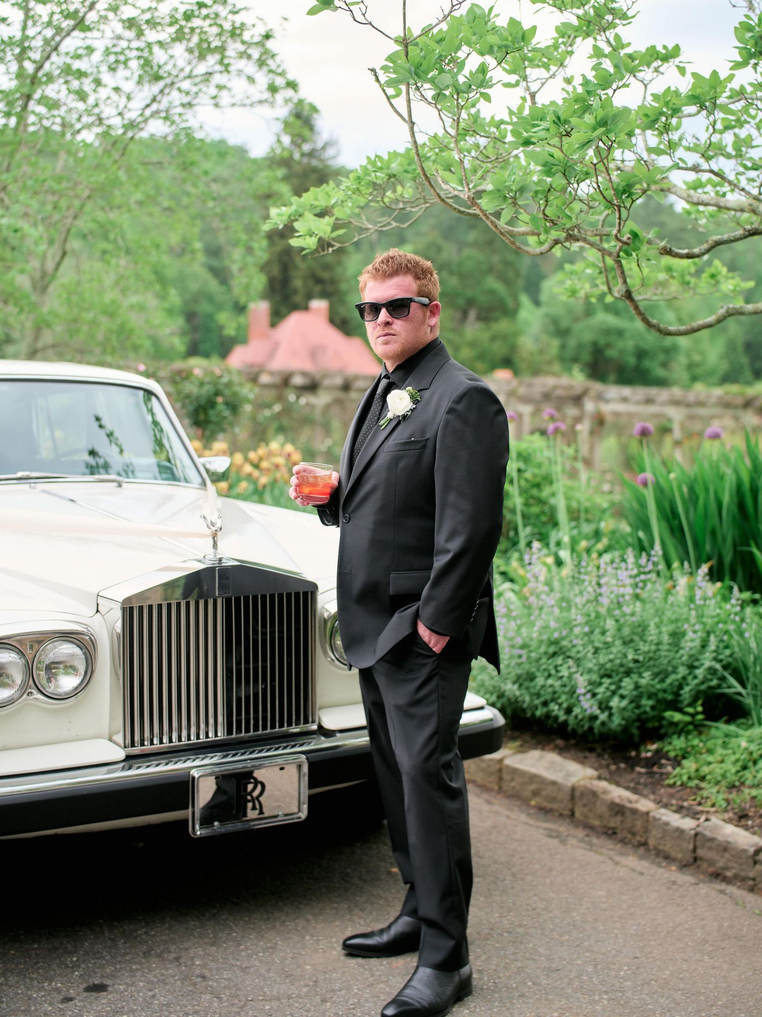 A classic white Rolls Royce luxury car parked in a garden setting with lush greenery in the background.