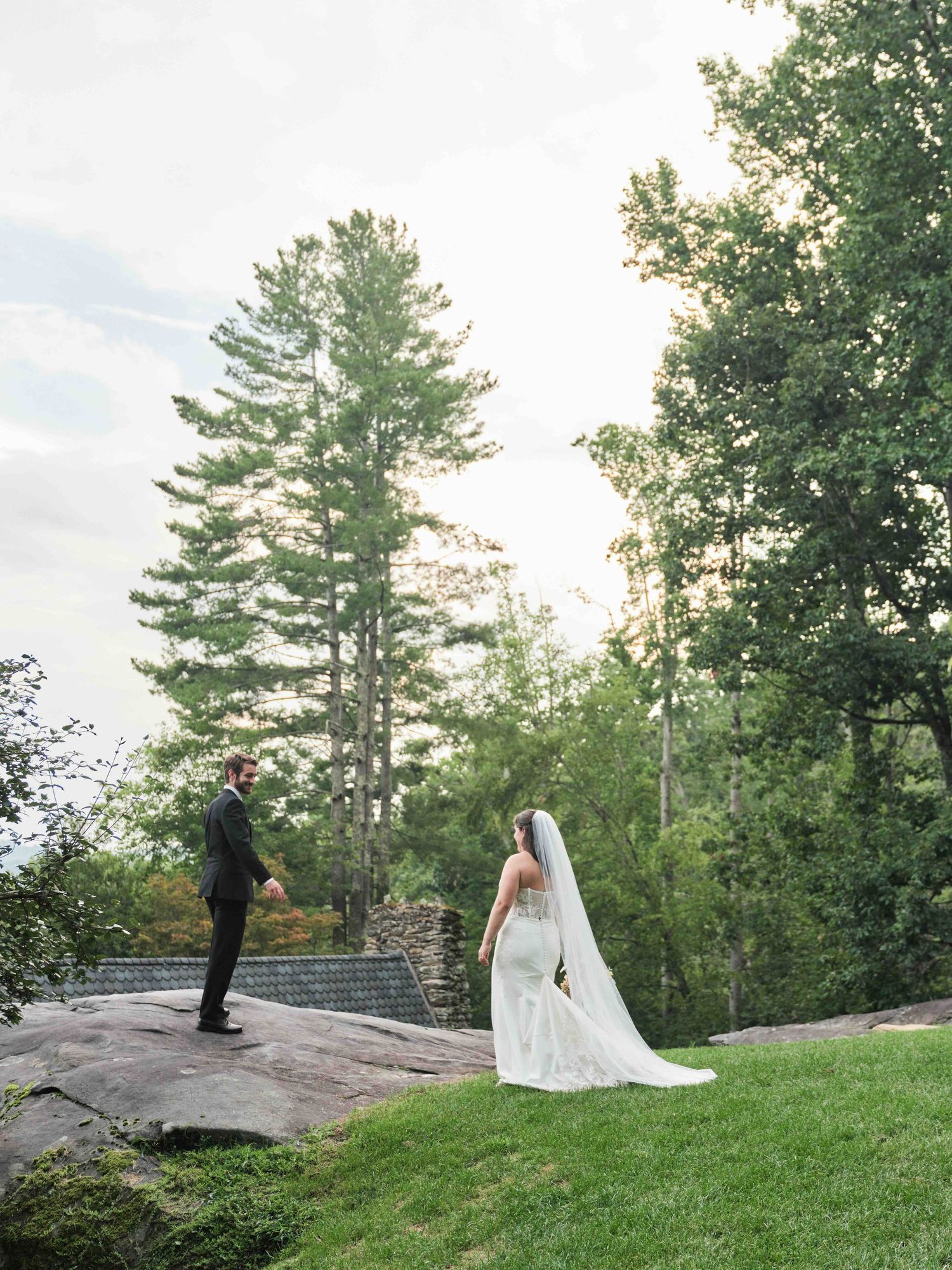 Bride in lace gown and veil faces groom on rocky outcrop surrounded by lush green trees at outdoor wedding.