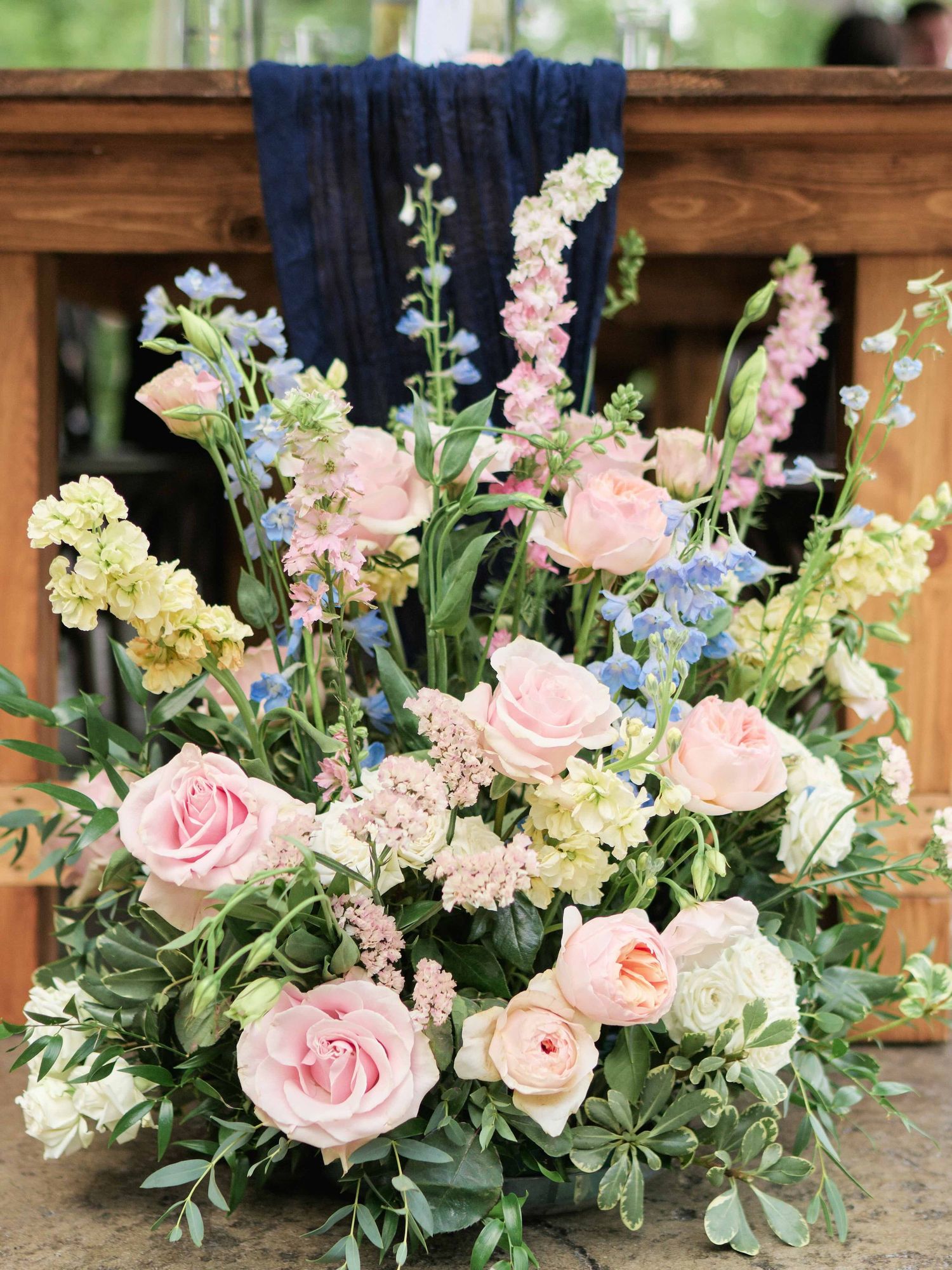 Lush floral arrangement featuring blush pink roses, blue delphiniums, and greenery displayed on a rustic wooden table.