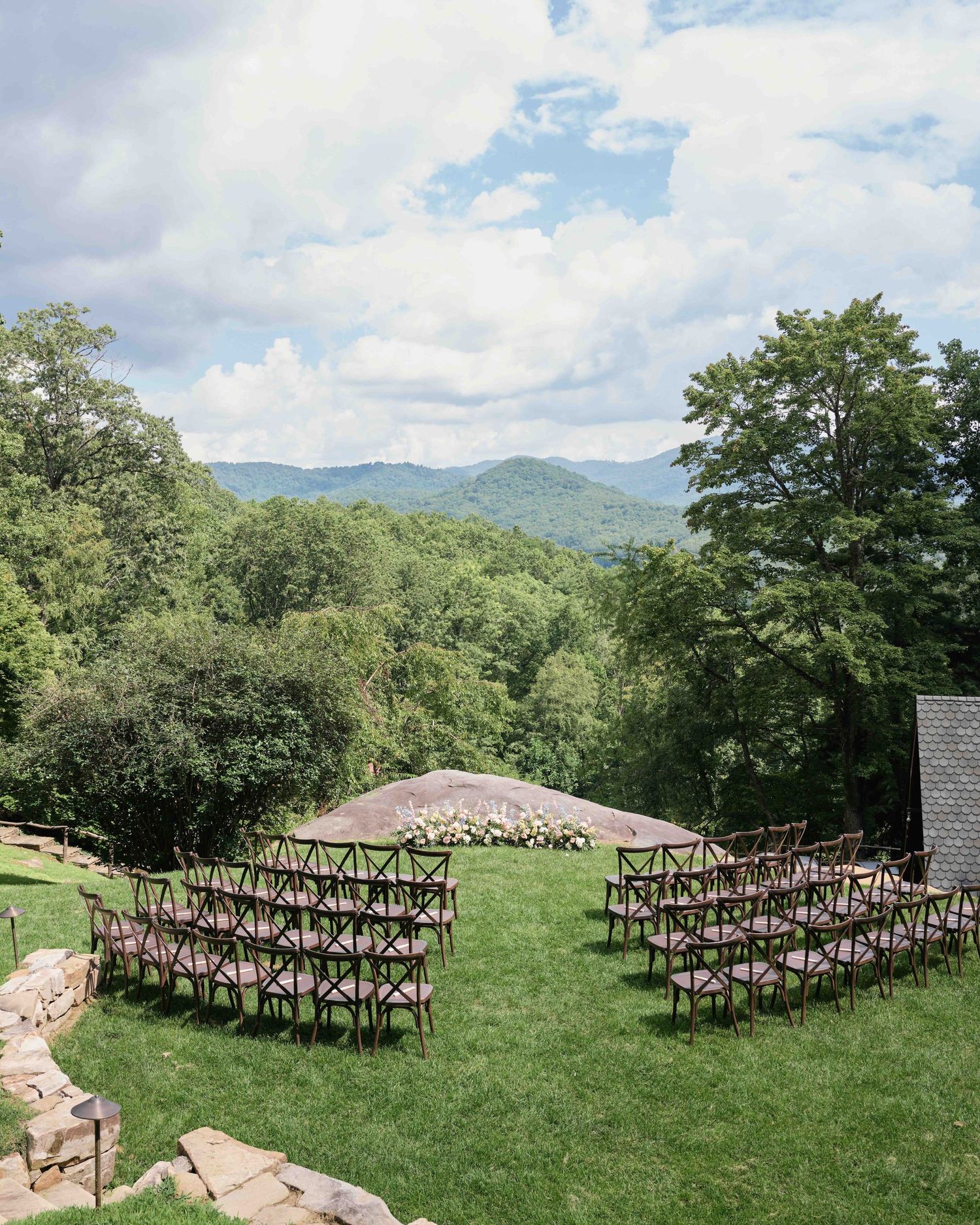 Outdoor wedding ceremony setup with wooden chairs on green lawn, mountain views and lush forest backdrop under cloudy sky.